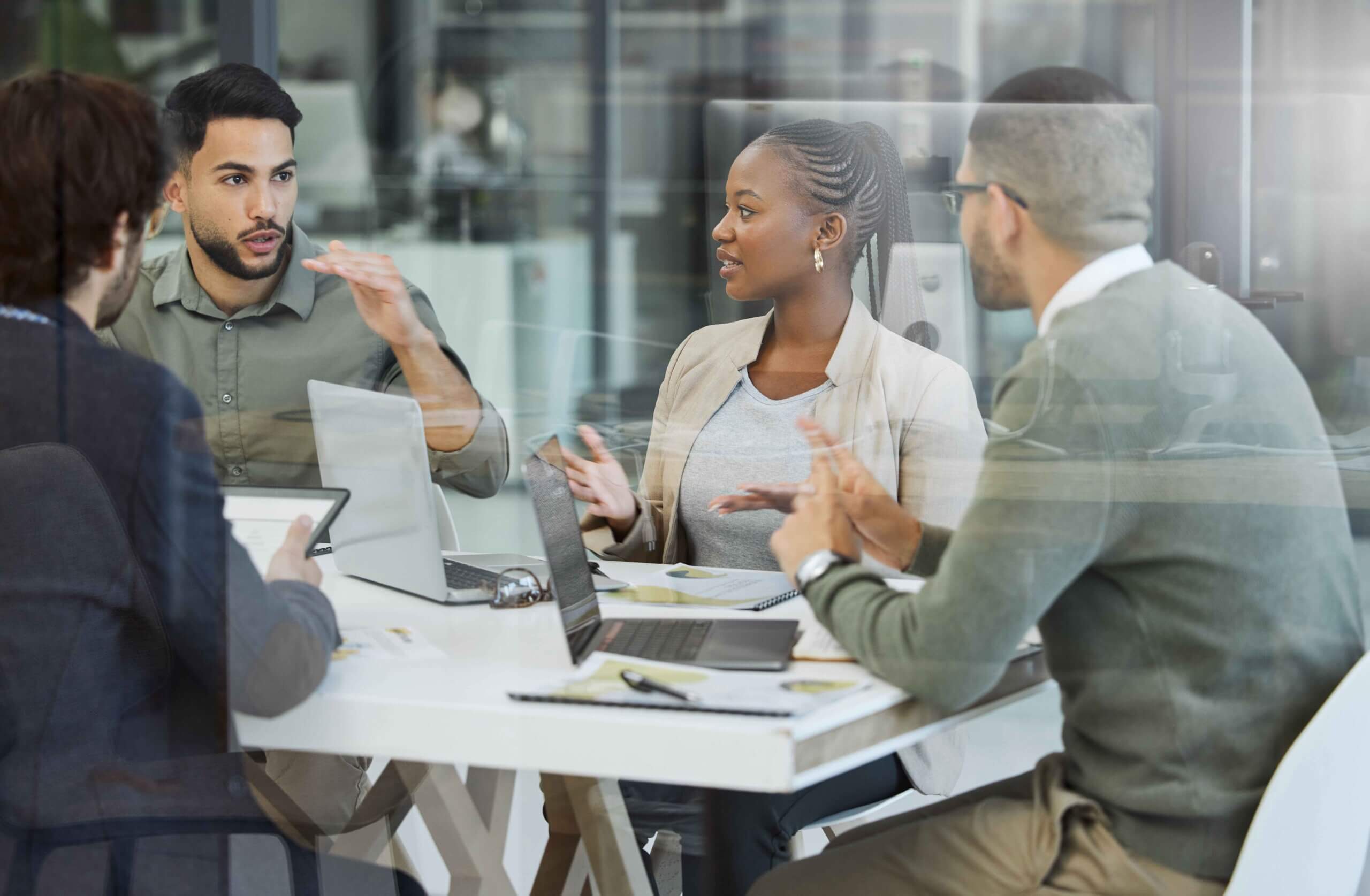 Four people sit around a table in an office, engaged in a discussion. Laptops and notebooks are on the table, and the group appears to be collaborating on a project or having a meeting.