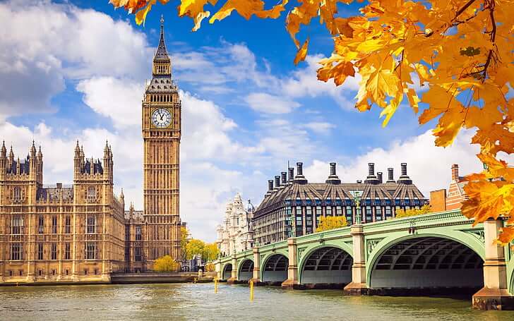 View of Big Ben and the Houses of Parliament beside the River Thames in London, with Westminster Bridge in the foreground and yellow autumn leaves framing the scene under a blue sky.