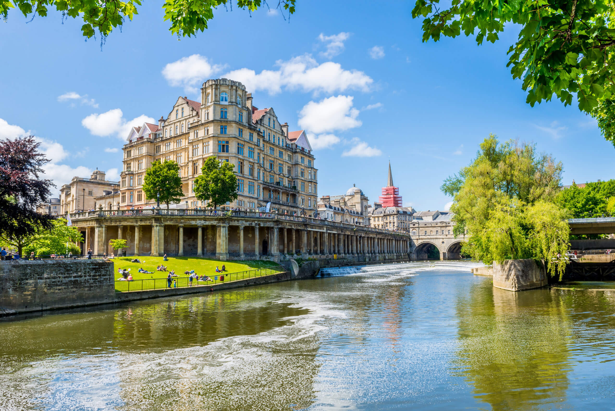 A grand historic building overlooks a river with green lawns and trees under a bright blue sky. People sit along the riverbank, and a stone bridge is visible in the background.