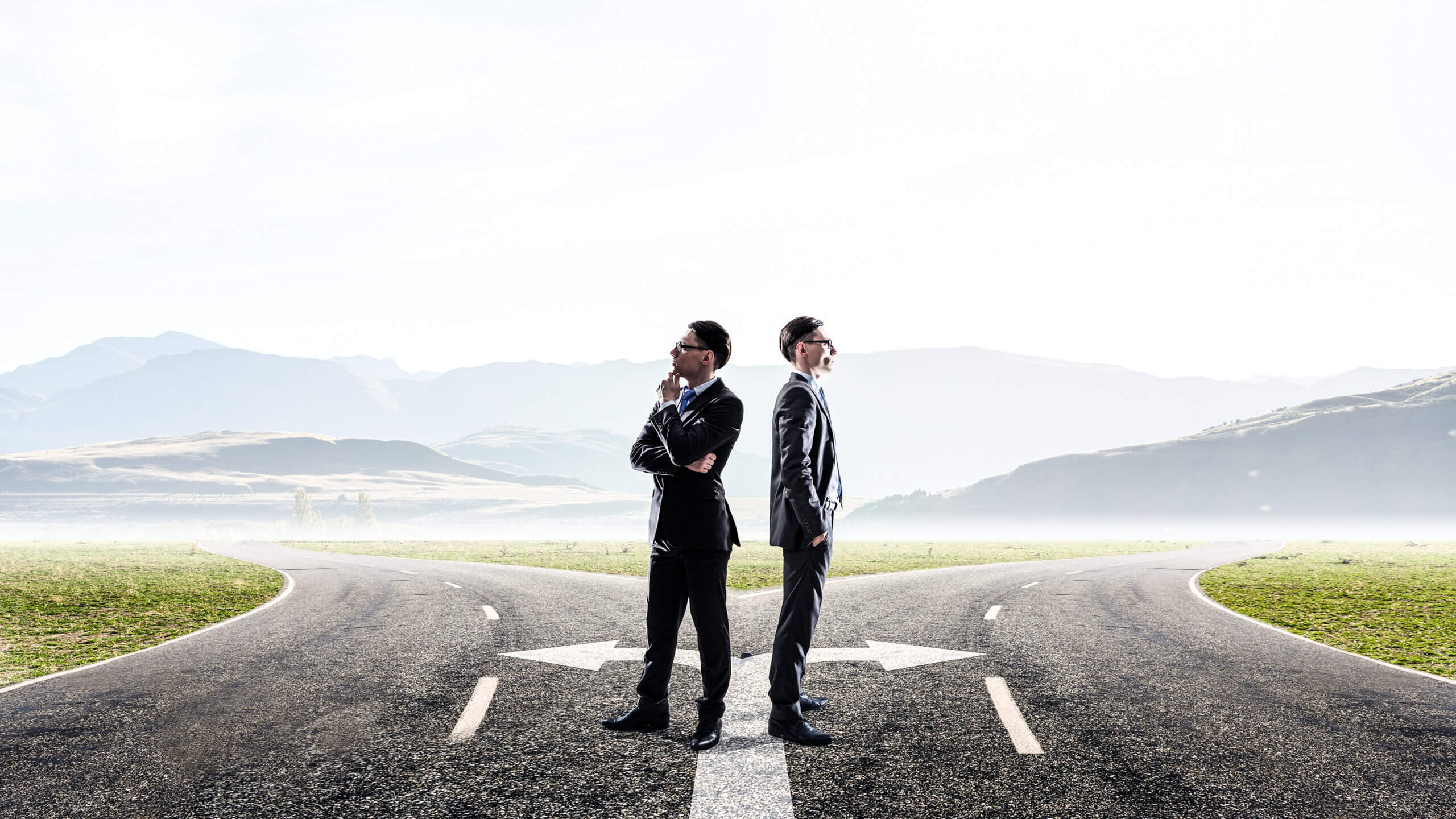 Two businessmen in suits stand back-to-back at a fork in the road, each facing opposite directions, symbolizing decision-making or choosing different paths. Mountains and fields are visible in the background.