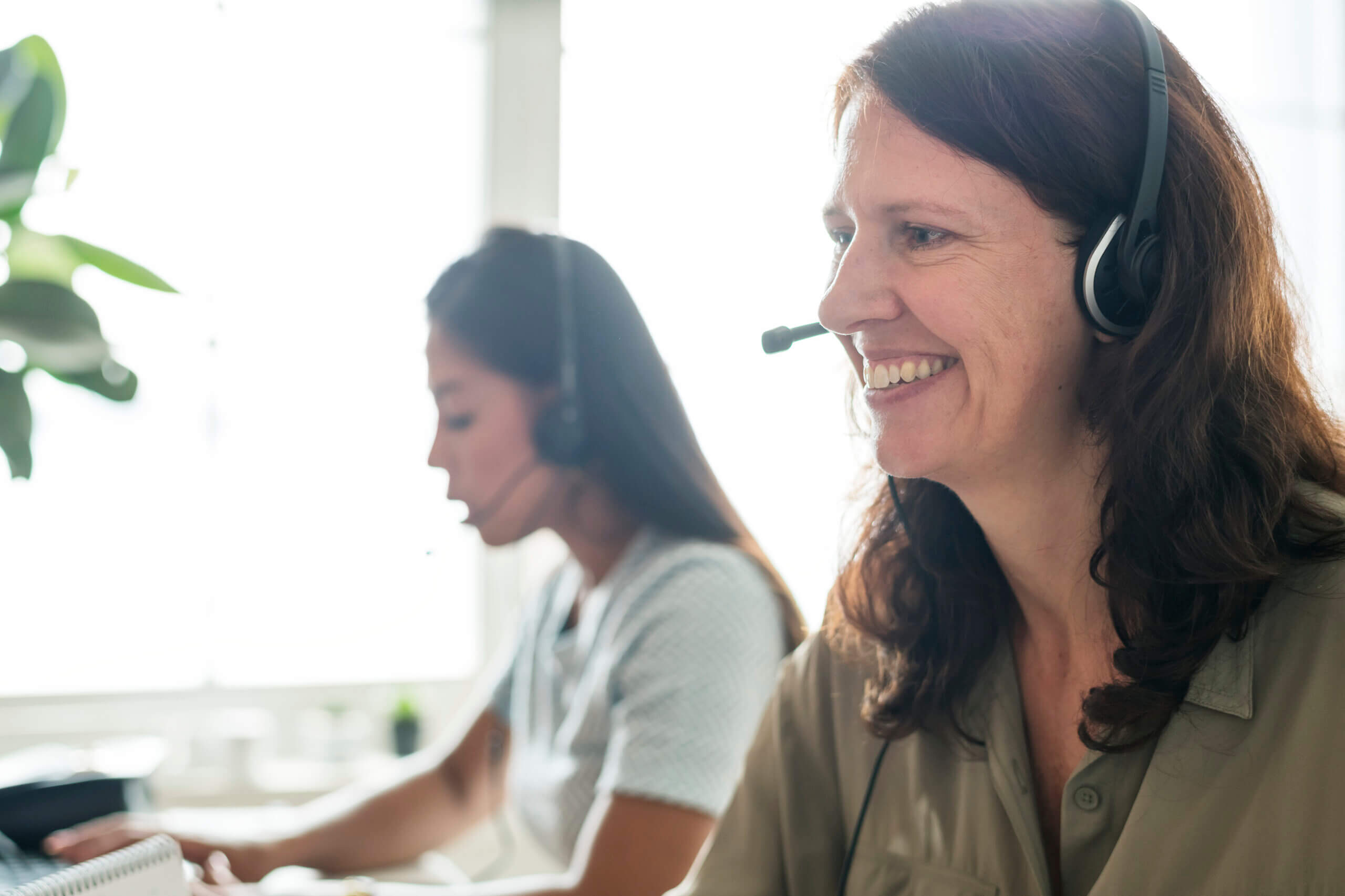 Two women wearing headsets work at computers in a bright office. The woman in the foreground is smiling, while the woman in the background appears focused on her screen.