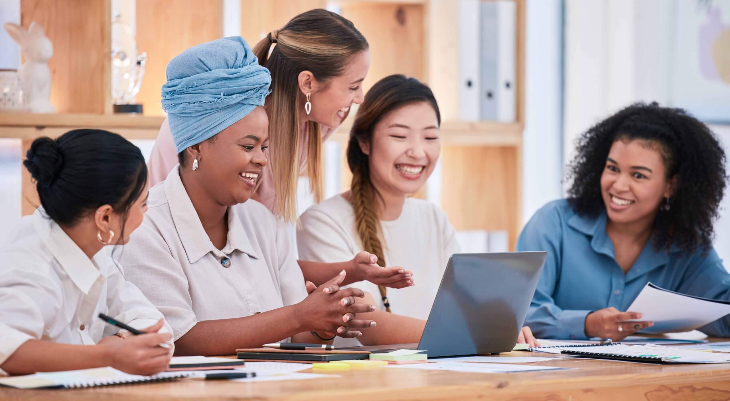 Five women sit around a table, smiling and talking while looking at a laptop. They have notebooks, pens, and papers in front of them, suggesting a collaborative meeting or discussion in a bright office space.