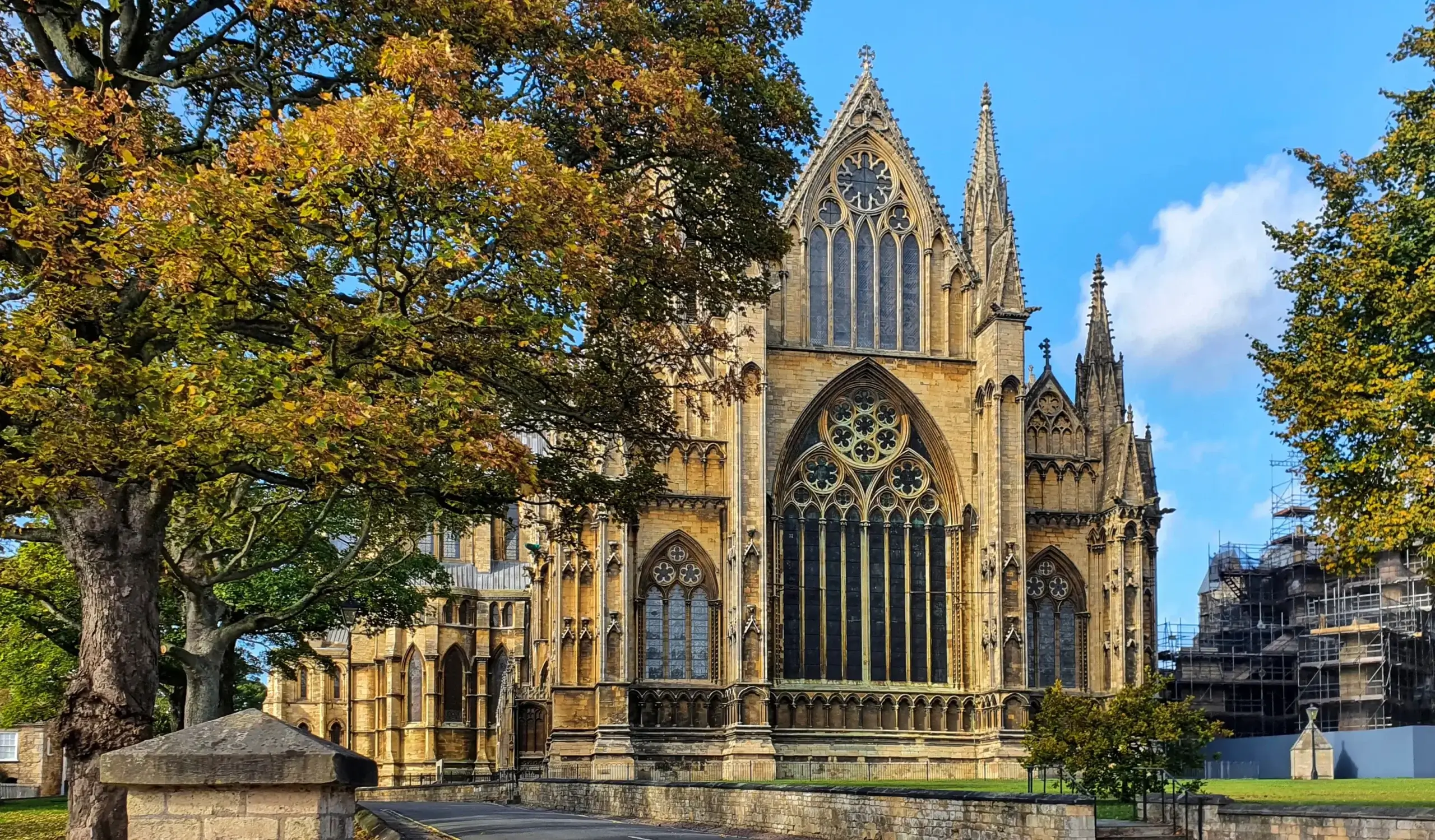 A large Gothic-style cathedral with pointed arches, tall stained glass windows, and ornate stonework, surrounded by autumn trees and a clear blue sky.