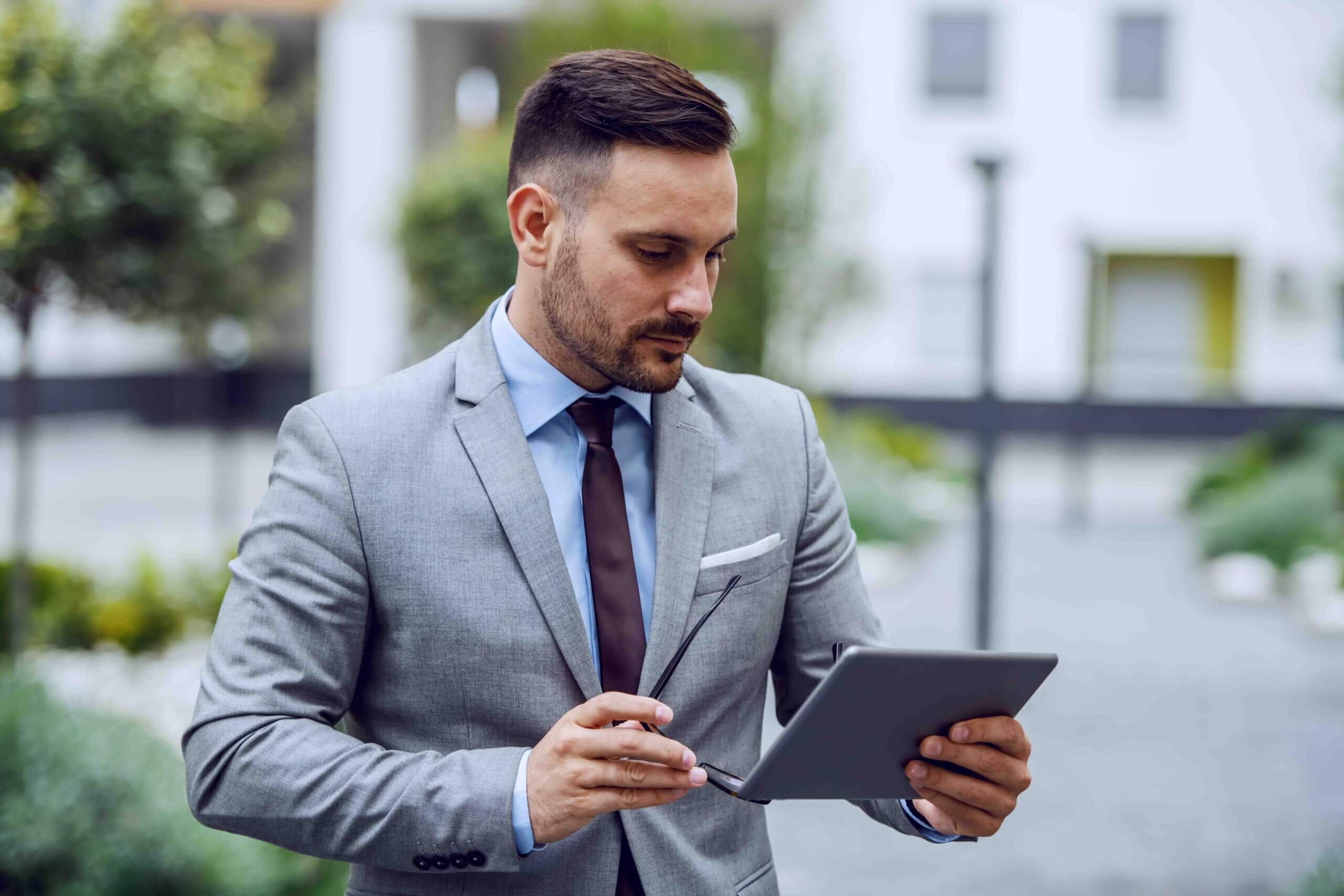 A man in a gray suit holding a tablet and eyeglasses stands outdoors, appearing focused as he looks at the screen. The background shows blurred greenery and buildings.