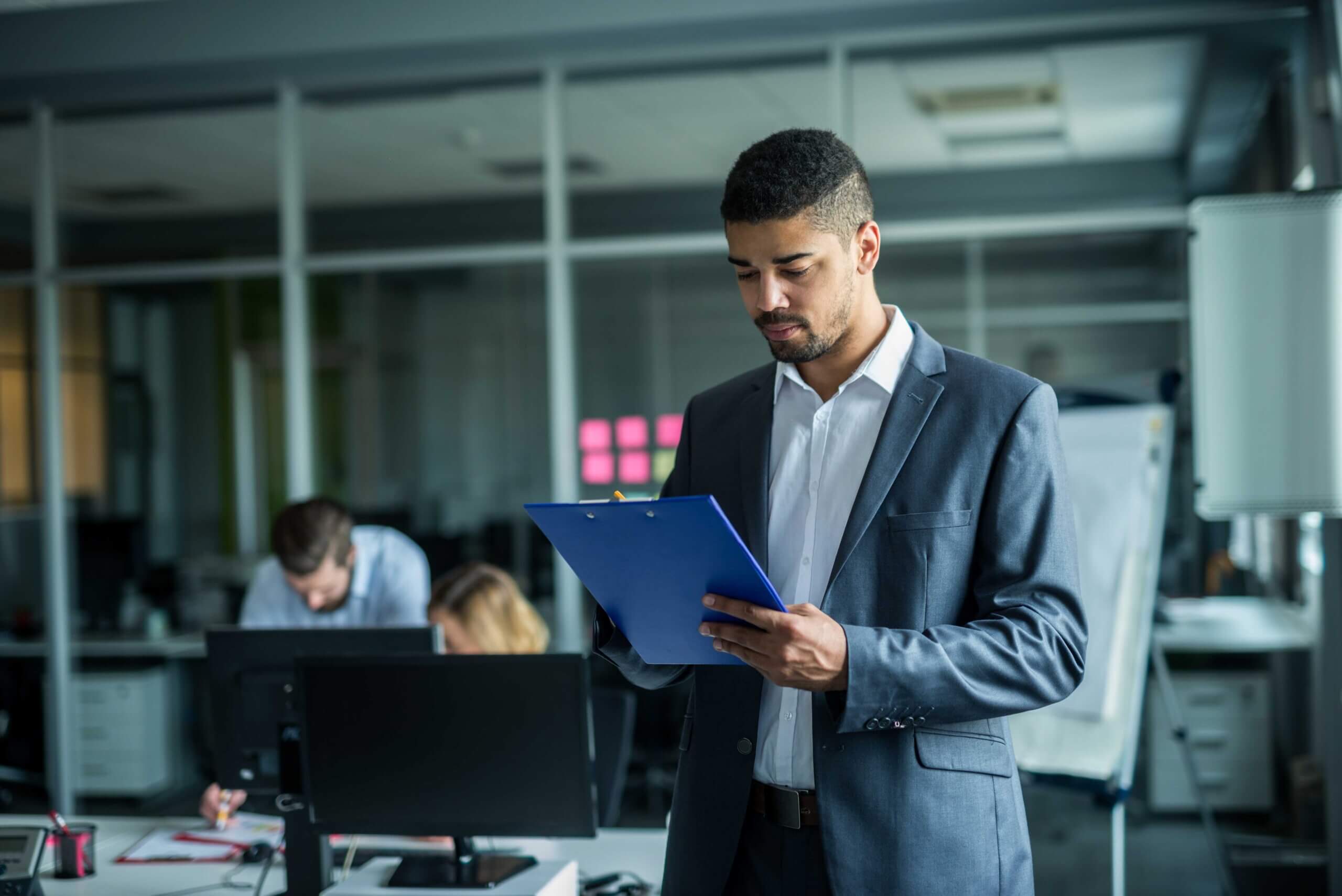 A man in a suit stands in an office, looking at a blue clipboard. Behind him, two colleagues work at desks with computers. The office has a modern, professional atmosphere.