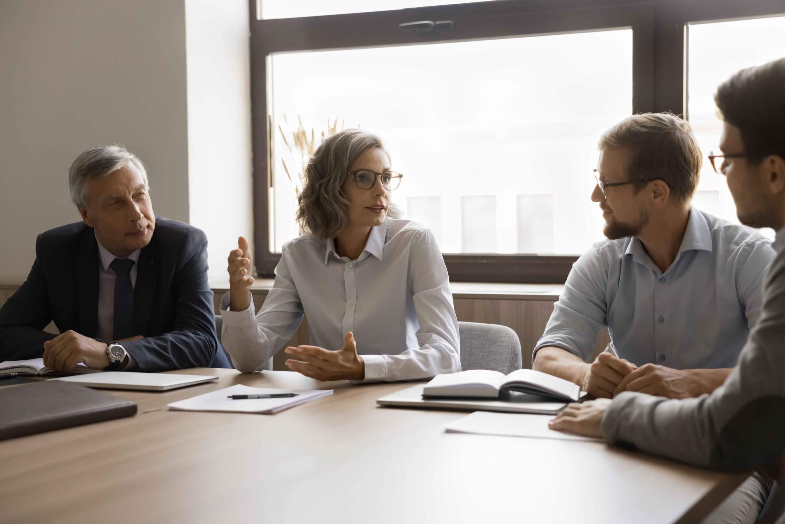 Four professionals sit at a table in an office, having a discussion. One woman gestures while speaking, and three men listen attentively. Papers and notebooks are on the table, and sunlight streams through a window behind them.