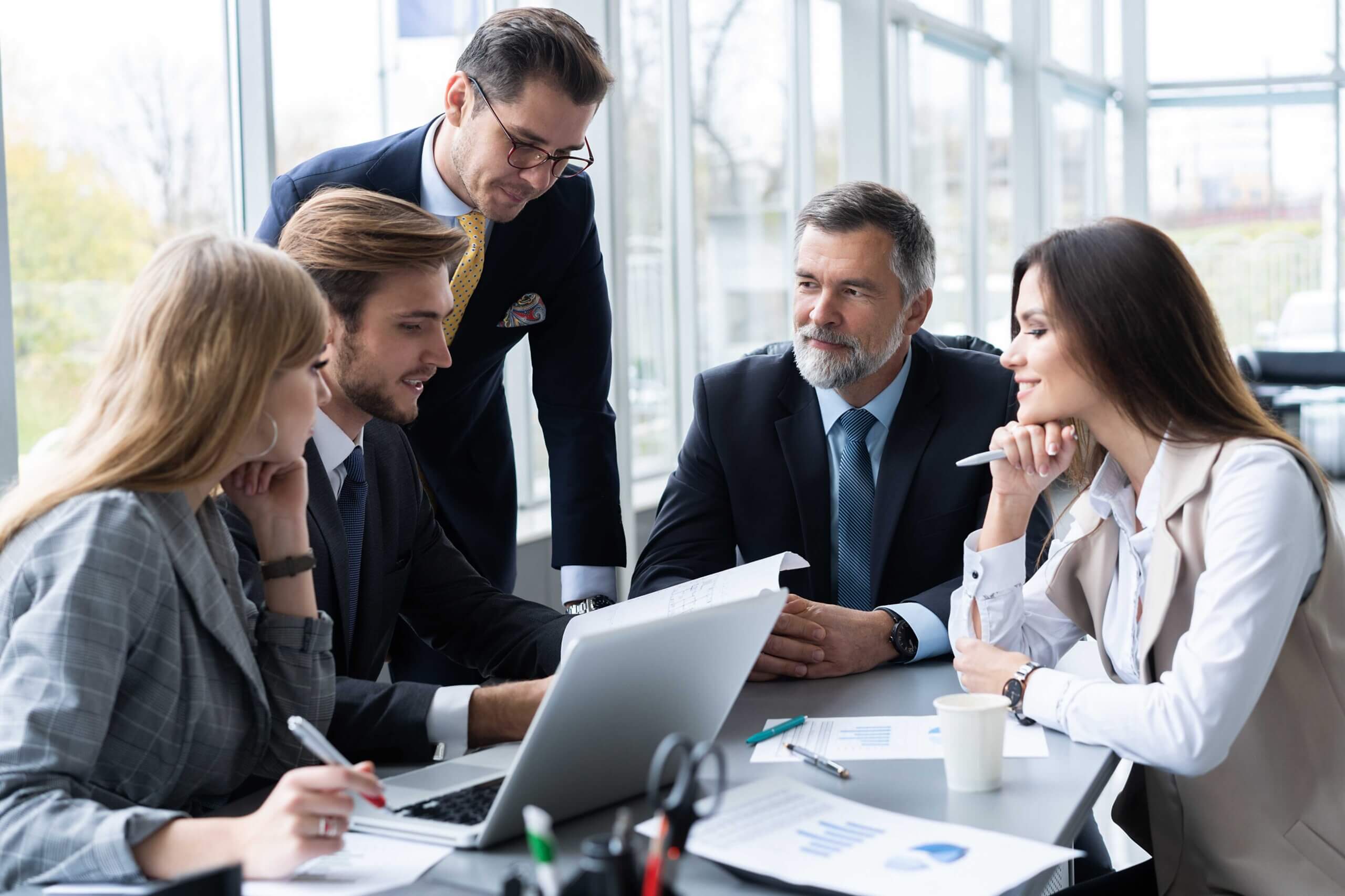Five business professionals are gathered around a table in an office, discussing documents and looking at a laptop, appearing engaged in a meeting or collaborative discussion.