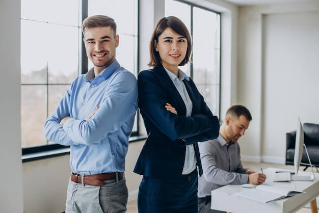 Two confident business professionals stand back-to-back with arms crossed, smiling, while a third person works at a desk in the background in a bright modern office.