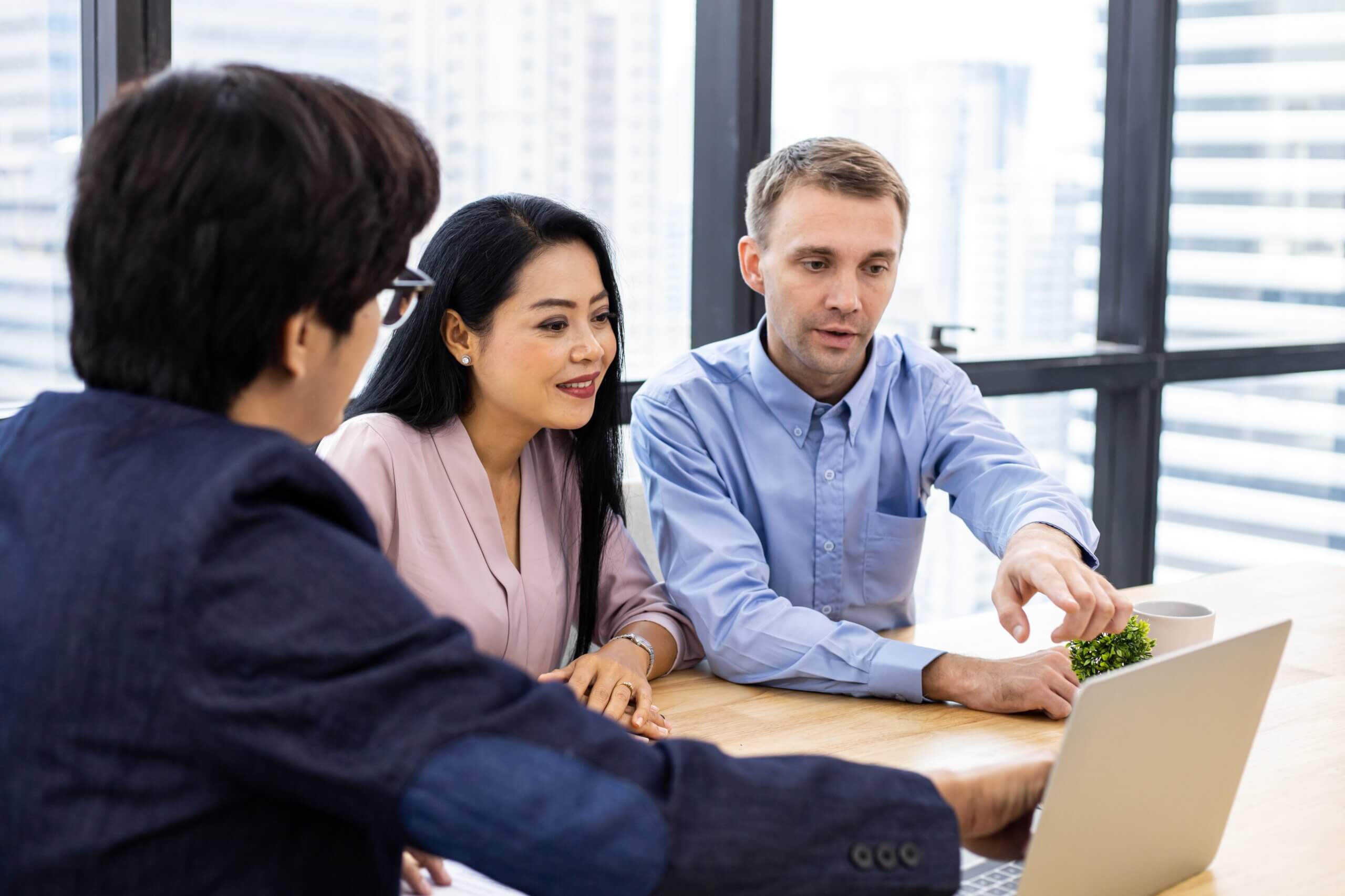 Three people sit at a table in an office, looking at a laptop. One man is pointing at the screen while discussing something with the other two, who are attentively listening. Large windows show a cityscape outside.
