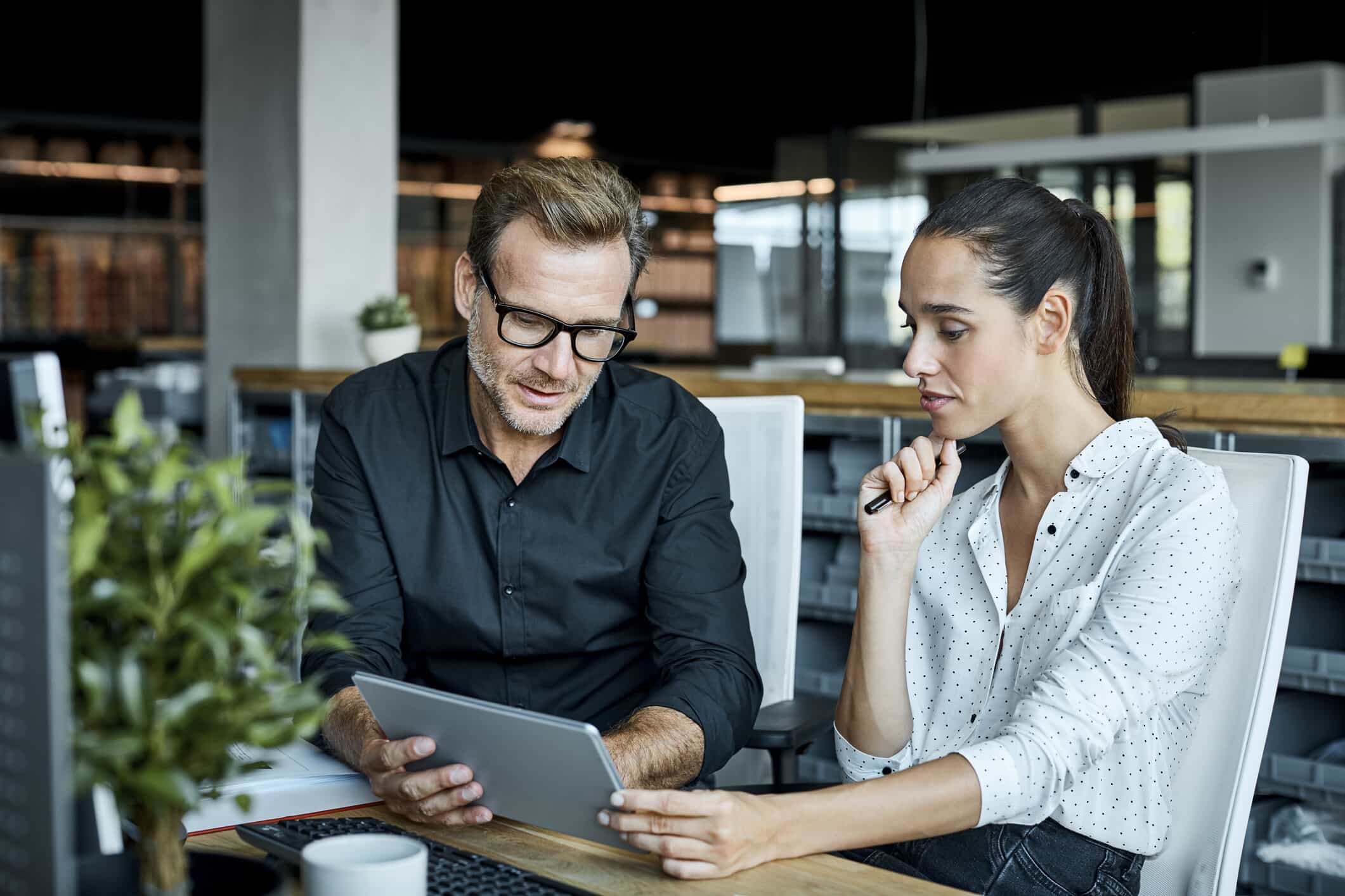 A man and a woman sit at a desk in a modern office, looking at a tablet together. The man wears glasses and a black shirt, while the woman wears a white blouse with black dots and holds a pen.