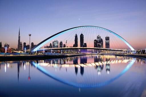 Modern arched bridge illuminated at dusk, spanning over calm water with the city skyline and skyscrapers in the background; the scene is reflected clearly in the water.