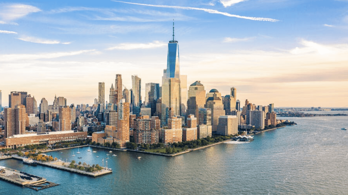 Aerial view of Lower Manhattan, New York City, featuring the tall One World Trade Center tower surrounded by skyscrapers, with the Hudson River in the foreground under a blue sky.