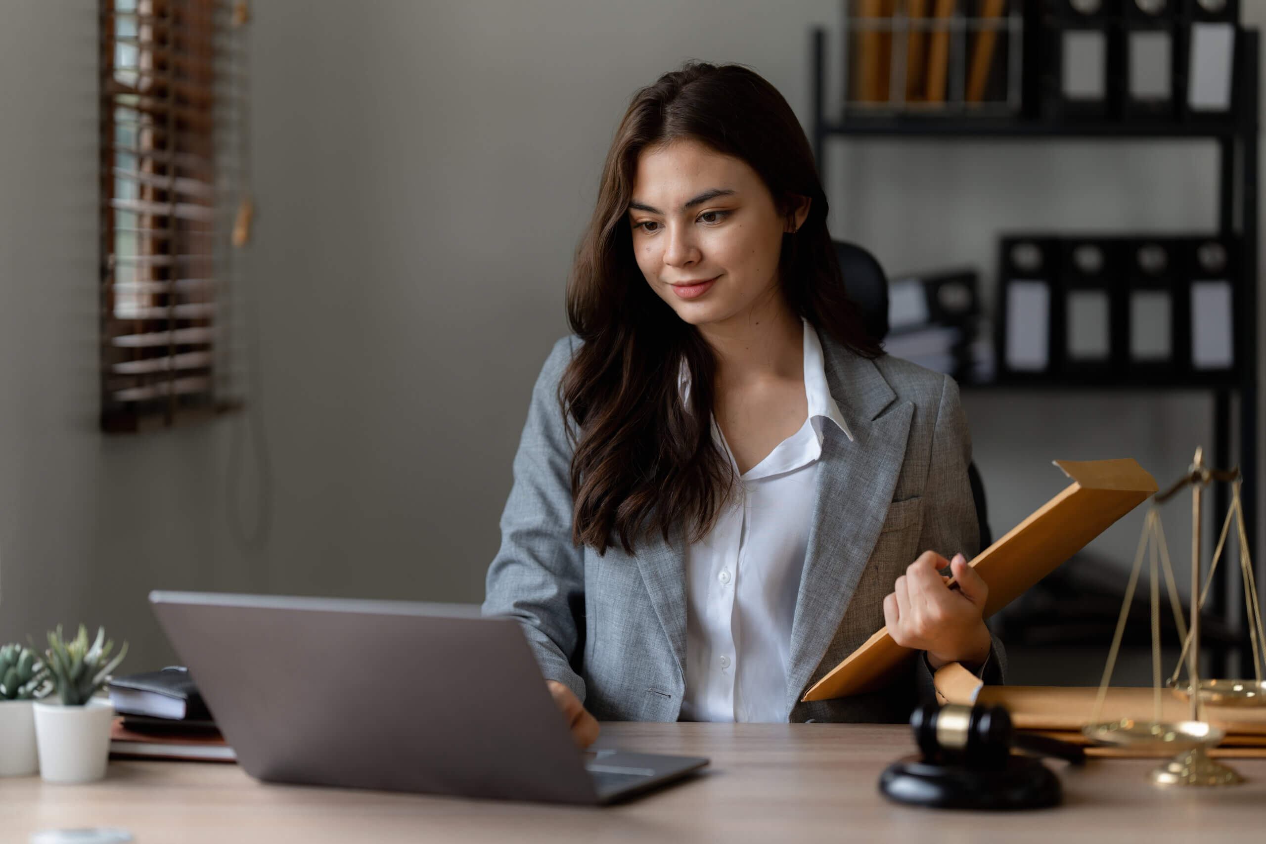 A woman in a gray blazer sits at a desk, holding a folder and looking at a laptop. Legal scales and a gavel are on the desk, suggesting a law office setting.