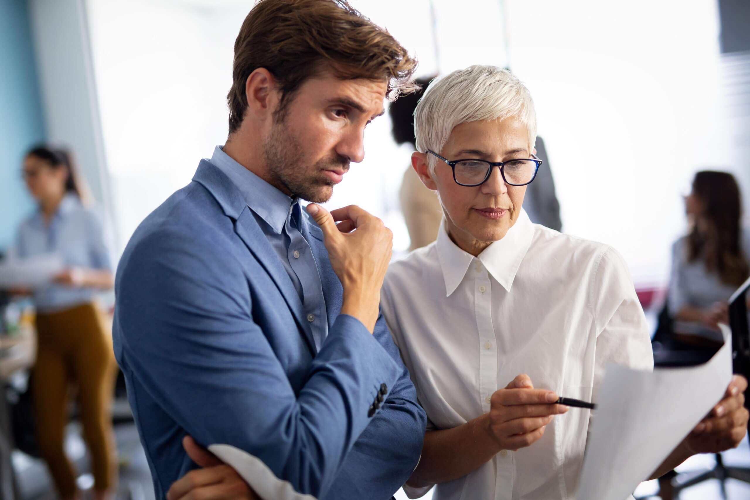 A man in a blue blazer and a woman with short gray hair and glasses discuss a document together in a bright office setting, both appearing focused and thoughtful.