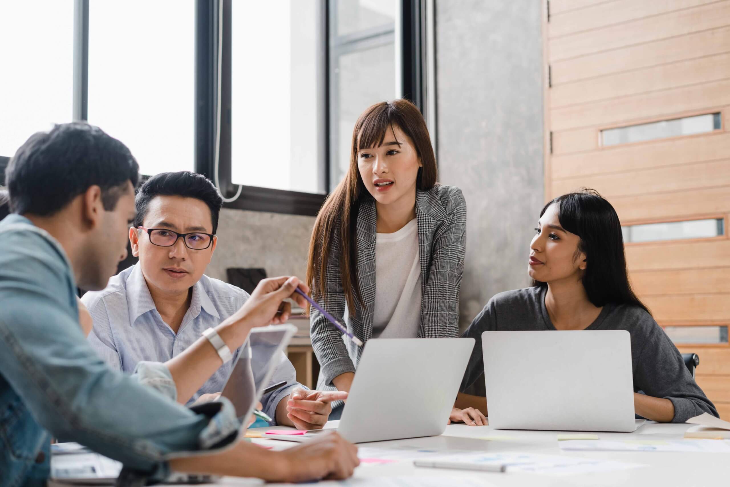Four colleagues sit and stand around a table with laptops, engaged in discussion. One person gestures while speaking, and the others listen attentively. Bright, modern office setting with large windows.