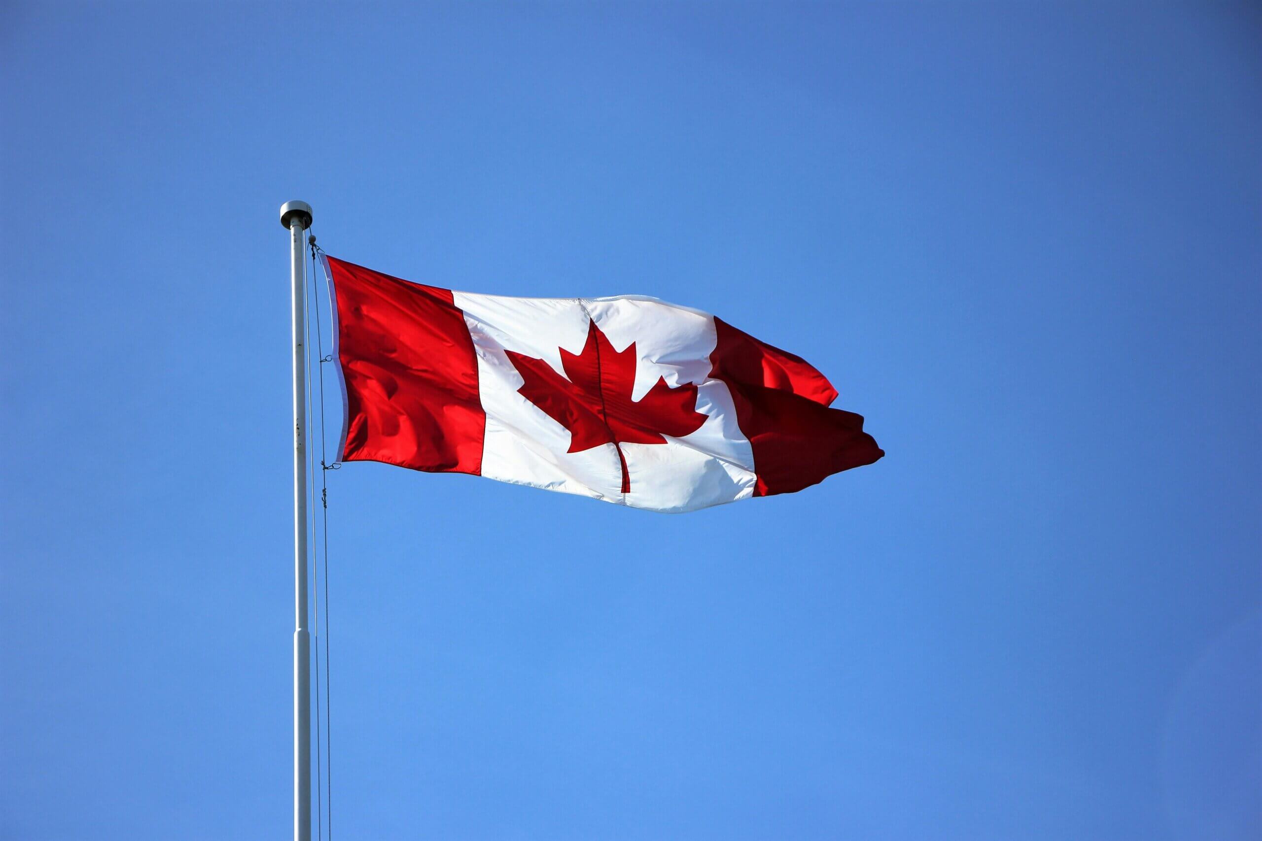 The Canadian flag with a red maple leaf in the center and red bars on each side is flying on a flagpole against a clear blue sky.