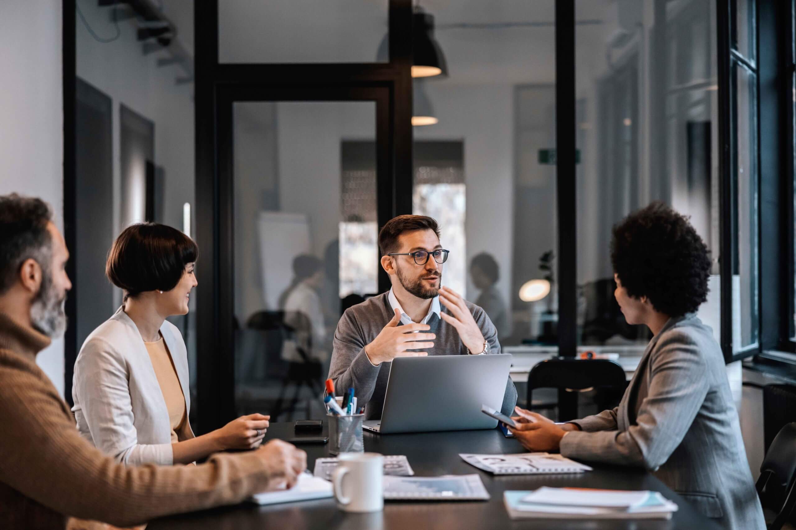 Four people in business attire sit around a conference table in a modern office, engaged in a discussion. One man speaks while others listen attentively. A laptop, documents, and coffee mugs are on the table.