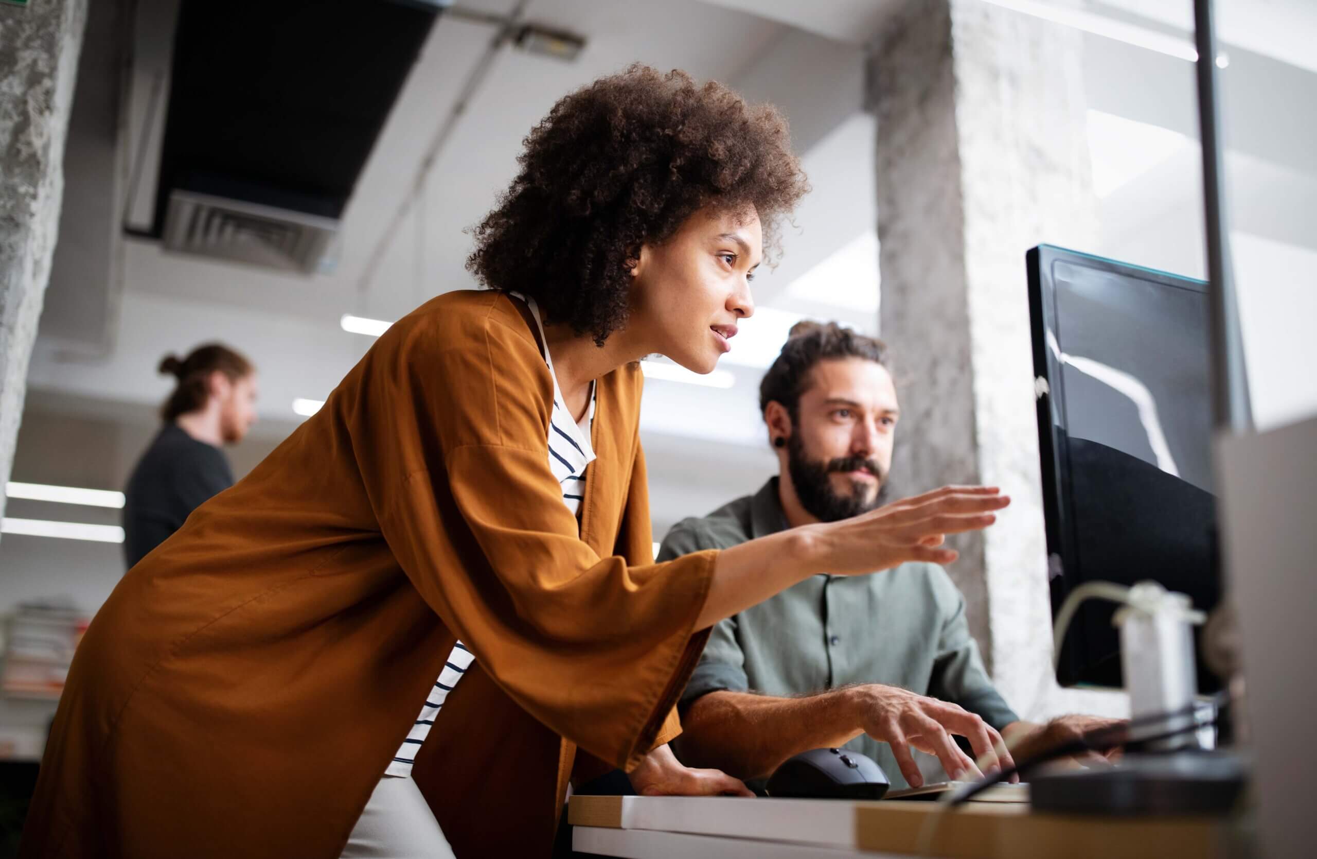 A woman leans over and points at a computer screen while talking to a seated man in a modern office. Another person is blurred in the background. Both appear to be collaborating on a project.