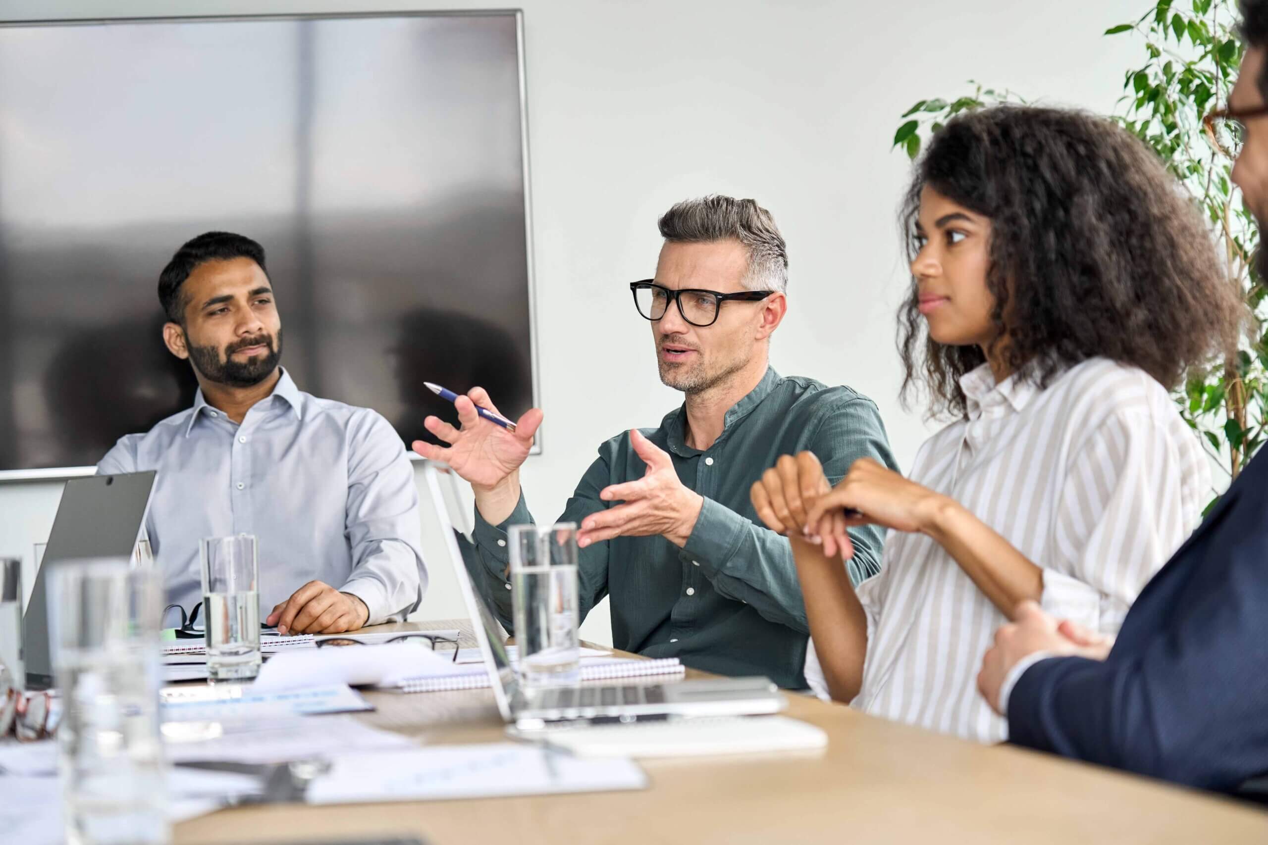 Four people sit at a conference table in a meeting room, with laptops, papers, and glasses of water. One man gestures as he speaks, while the others listen attentively. A large monitor and plant are in the background.