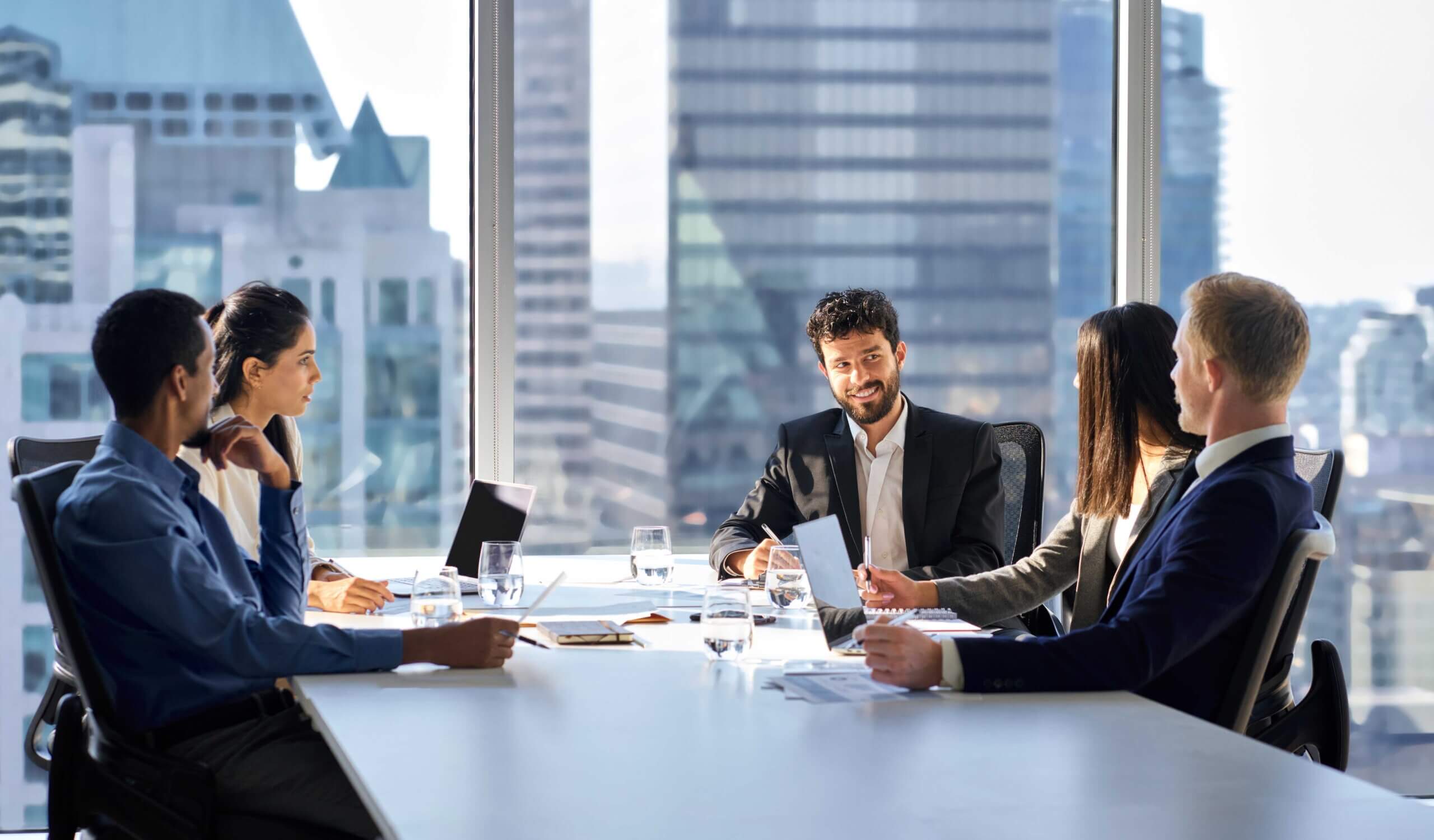 Five business professionals in formal attire sit around a conference table in a modern office with large windows, engaged in a meeting. City buildings are visible outside. Laptops, papers, and glasses of water are on the table.