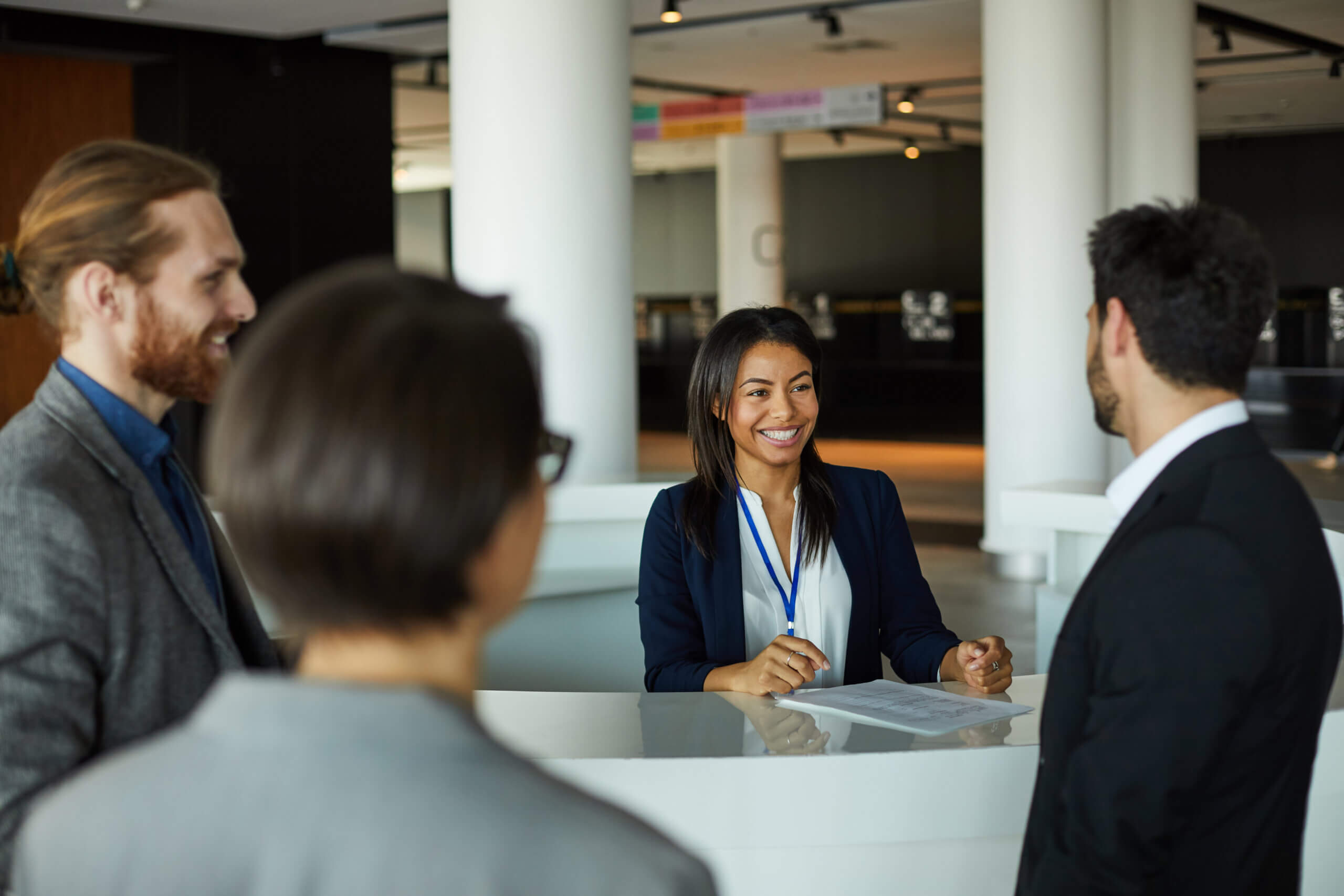 A smiling woman stands behind a reception desk, talking to three people in business attire in a modern office or lobby setting. She holds a pen and paper, while the others face her, engaged in conversation.