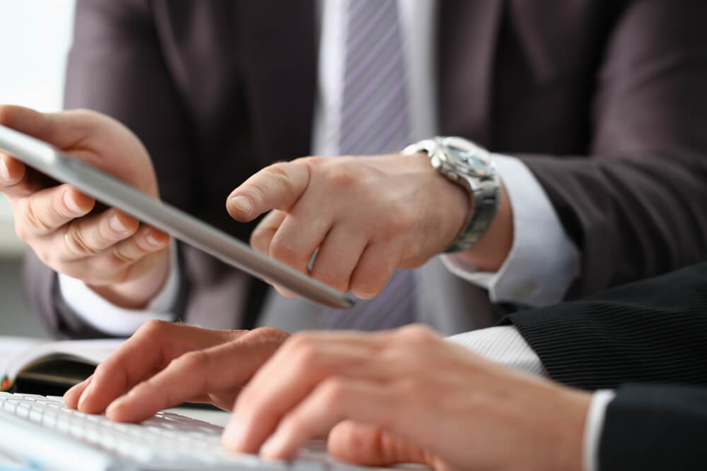 Two people in business attire are working together; one is typing on a keyboard while the other holds a tablet and points at its screen, suggesting a discussion or collaboration on a project.
