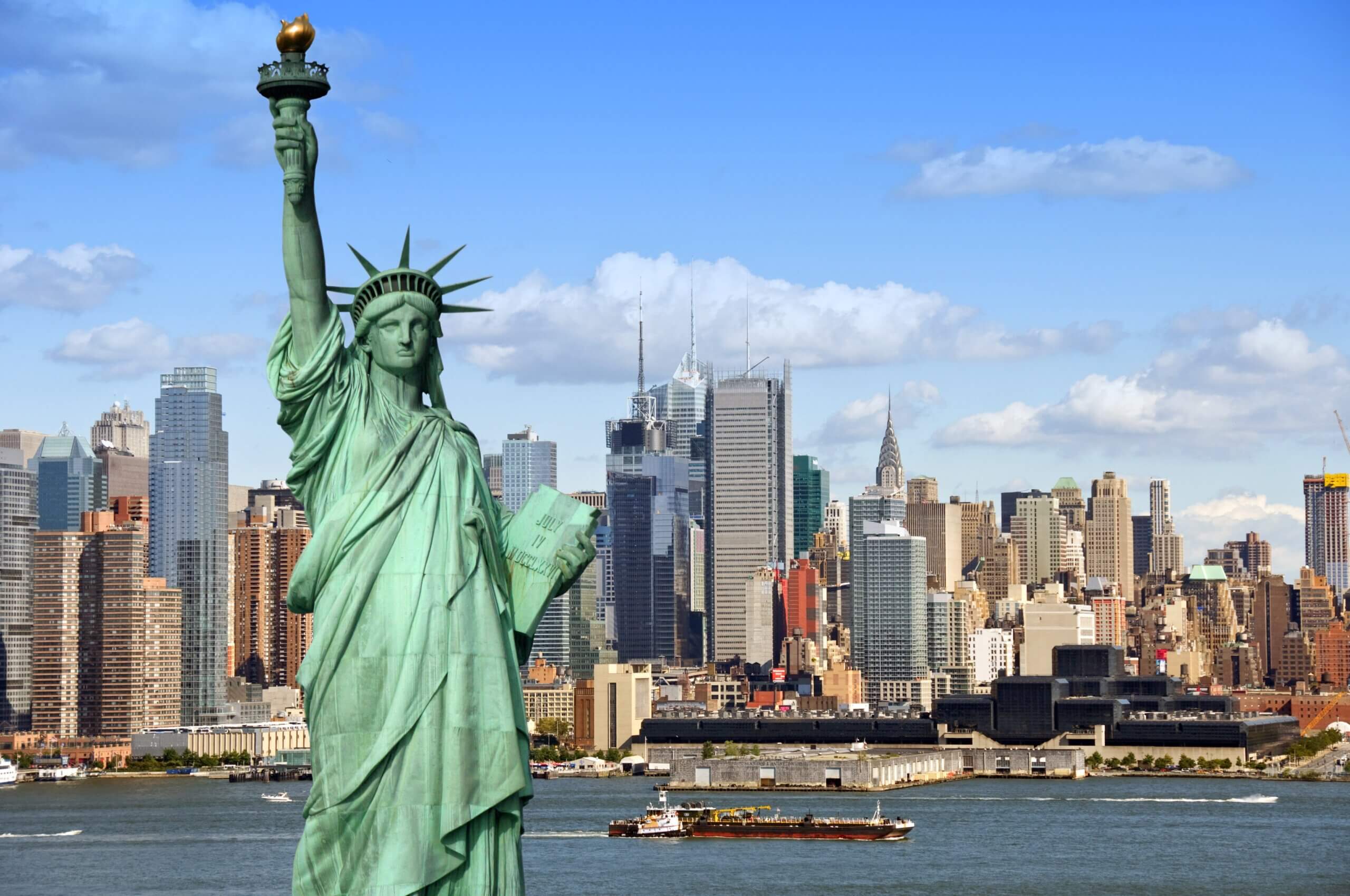 The Statue of Liberty stands in the foreground with the New York City skyline and skyscrapers in the background, seen across the water on a clear, sunny day.