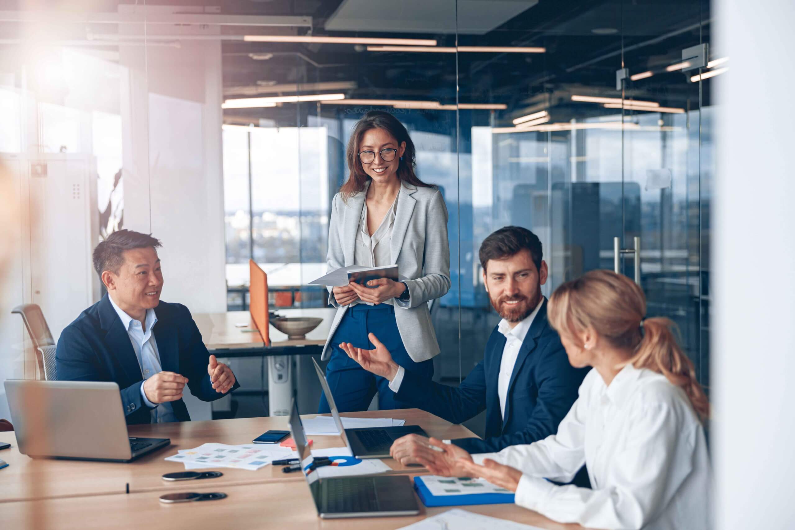Four business professionals, three seated and one standing, have a discussion in a modern office. Laptops, papers, and charts are on the table, and the group appears to be collaborating and smiling.