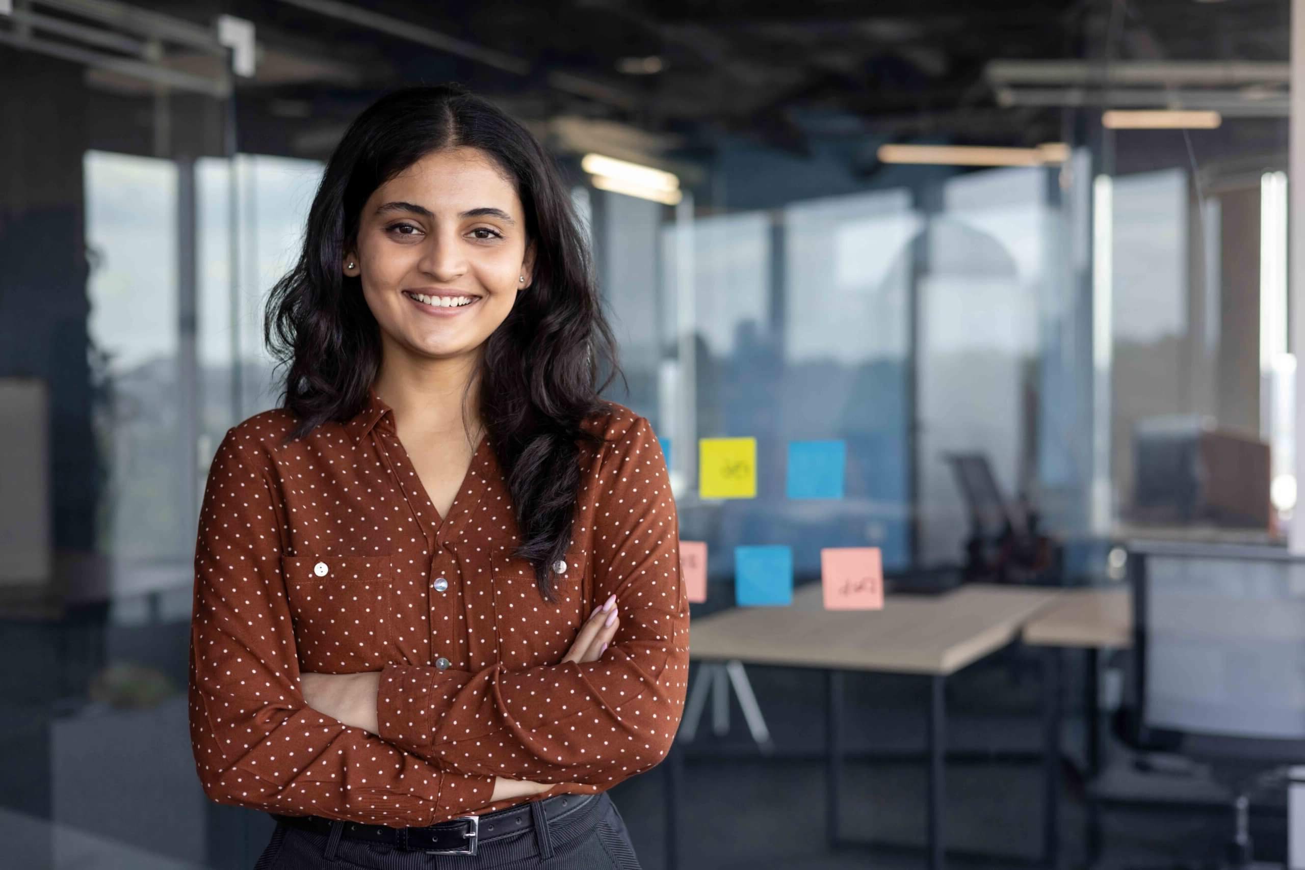 A woman with long dark hair, wearing a brown polka-dot blouse, stands smiling with arms crossed in a modern office with glass walls and colorful sticky notes on a window behind her.