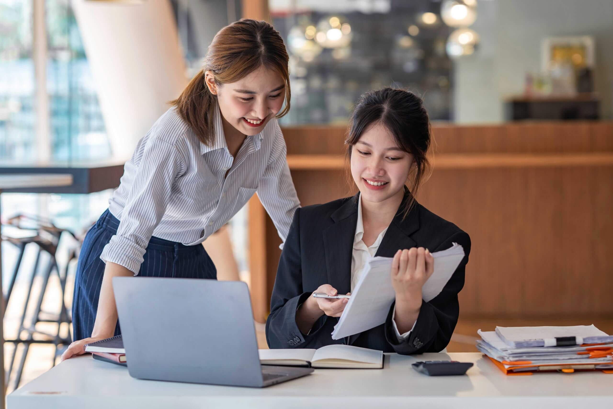 Two women working together at a desk with a laptop and documents. One woman is sitting, holding and reviewing papers, while the other stands beside her, smiling and looking at the documents.