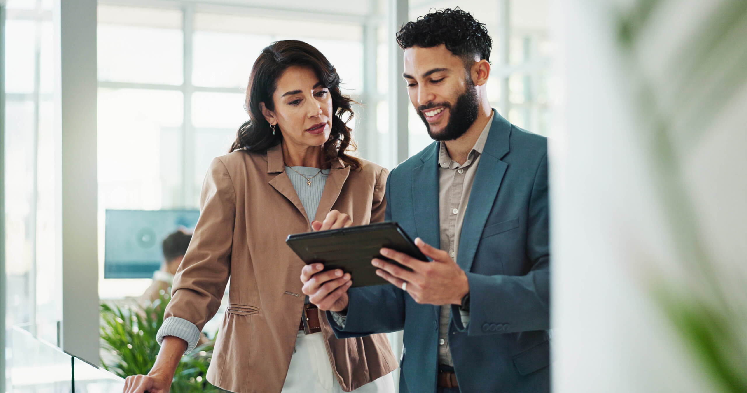 A woman and a man in business attire stand indoors, looking at a tablet together and discussing something. They appear to be in a modern office setting with large windows and natural light.