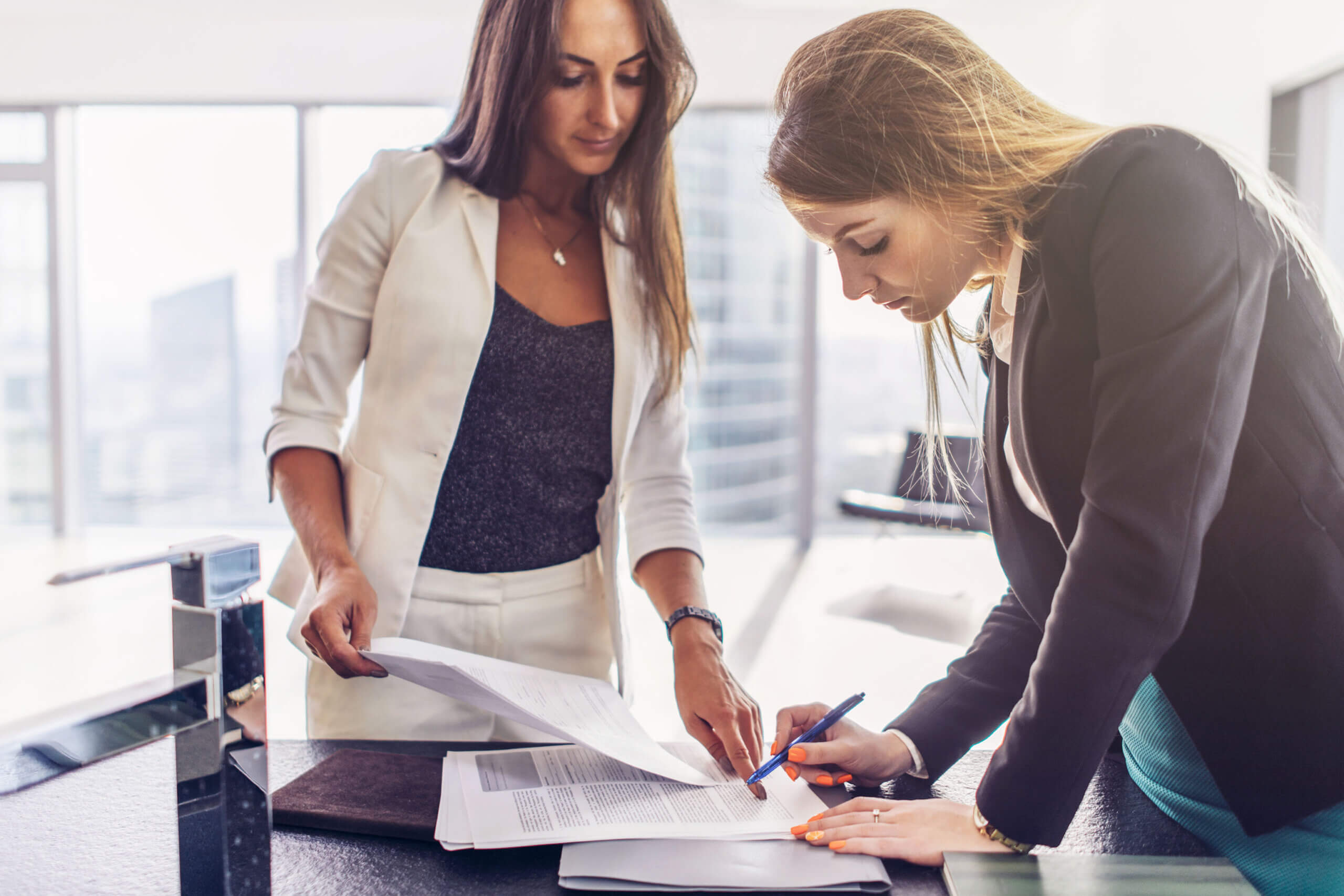 Two women in business attire review and sign documents at a desk in a bright office. One woman stands, holding papers, while the other bends over to write with a pen.