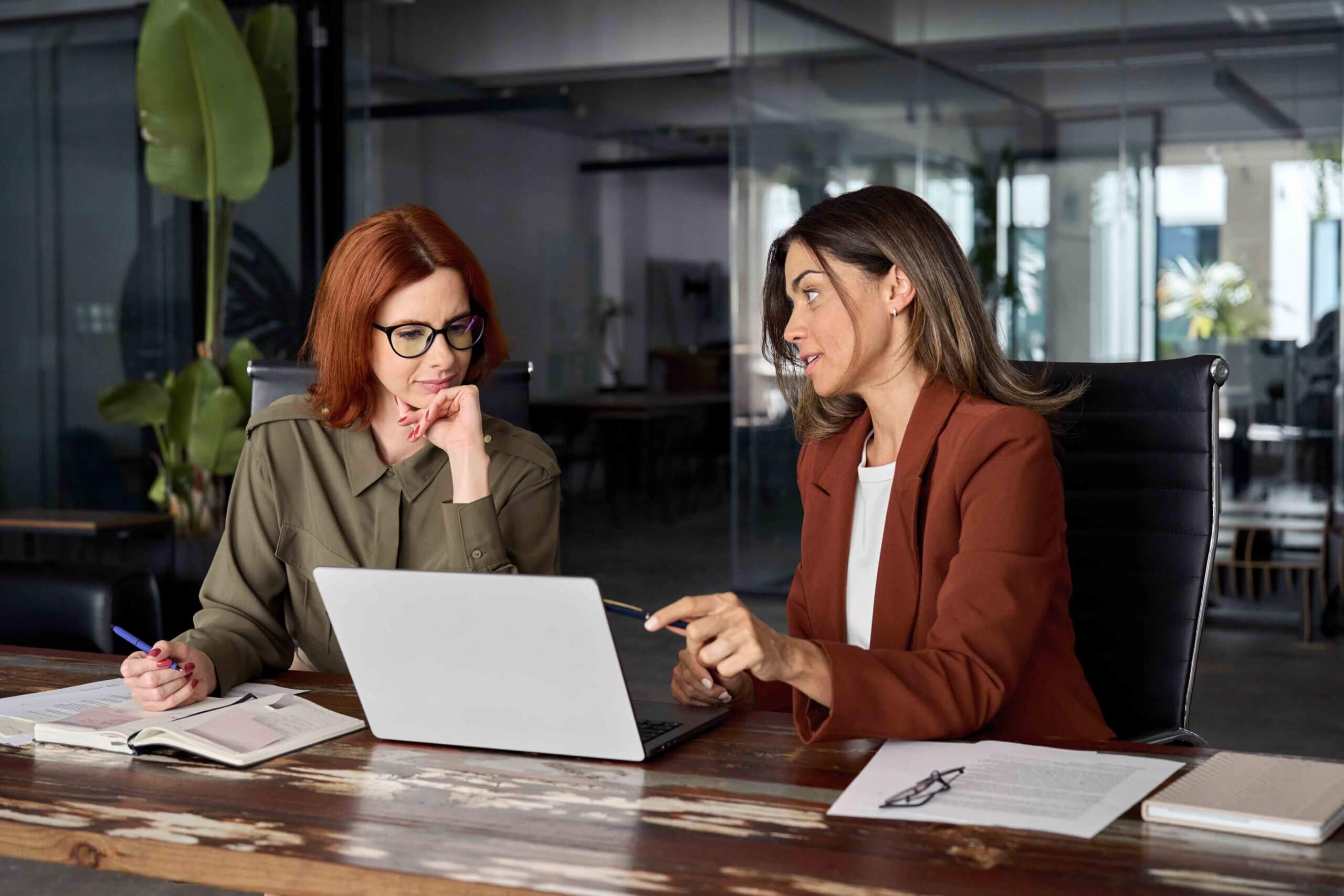 Two women sit at a table in an office, discussing something while looking at a laptop. One woman gestures toward the screen with a pen; the other listens thoughtfully. Papers and notebooks are on the table.