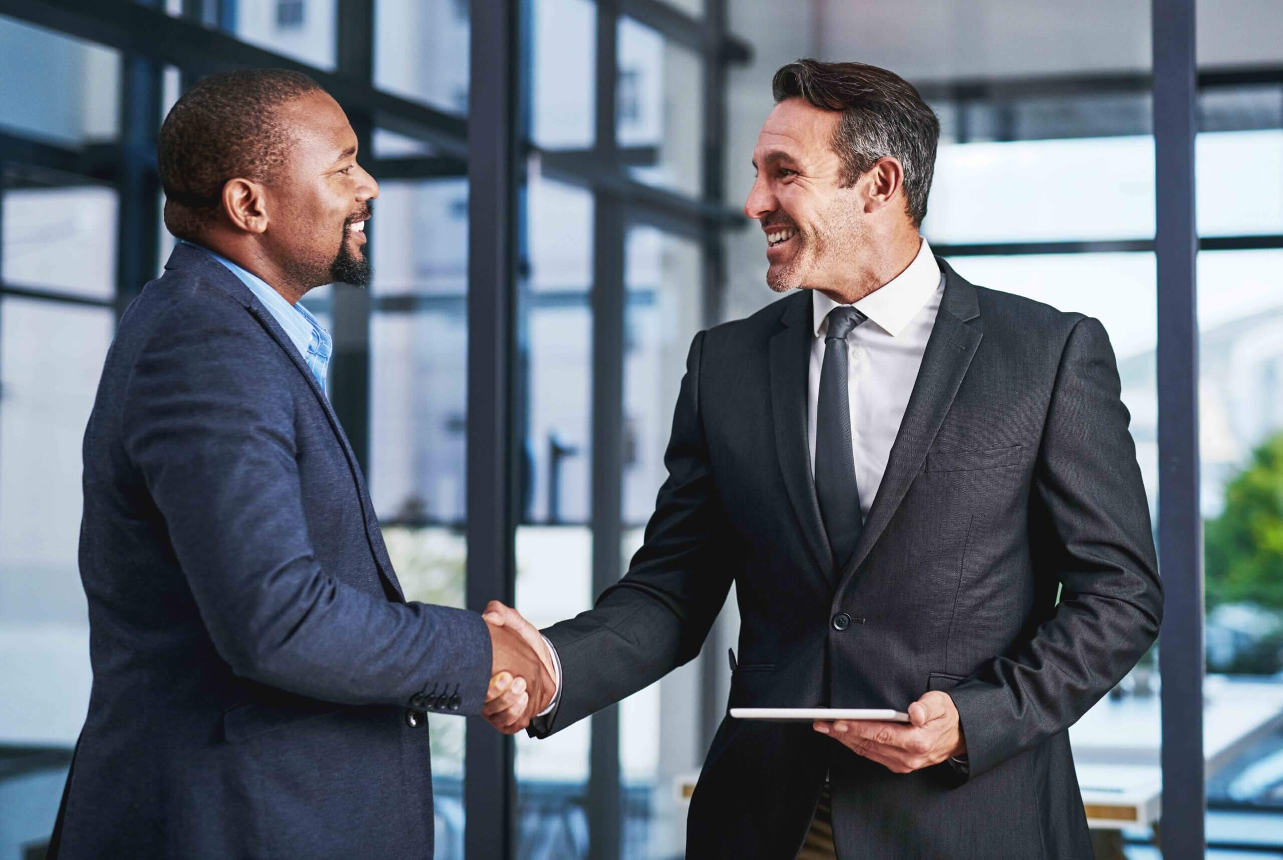 Two businessmen in suits smile and shake hands in an office setting, with one holding a tablet. Large windows and glass walls are visible in the background.
