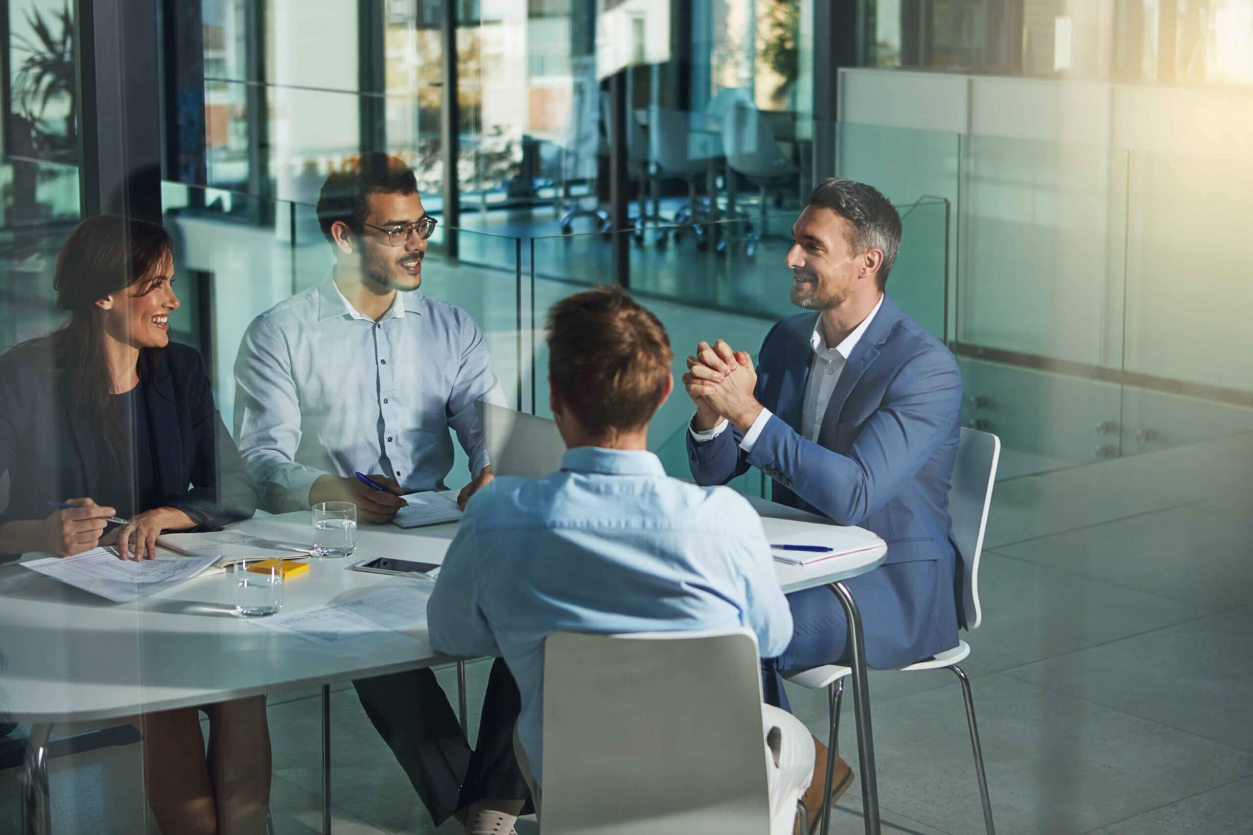 Four business professionals sit at a round table in a modern office, engaged in a discussion. Papers, pens, and glasses of water are on the table. The atmosphere appears collaborative and positive.