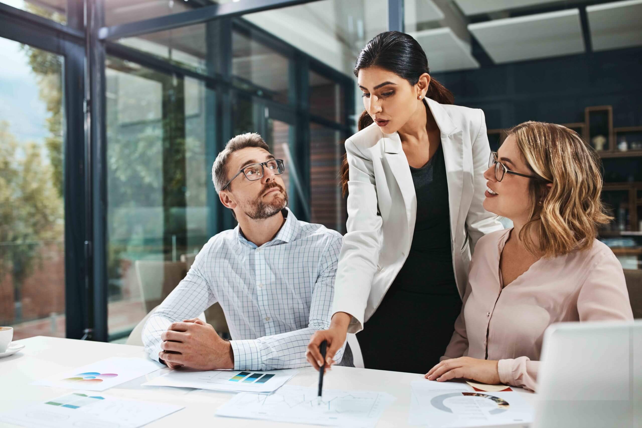 Three business professionals in an office discuss documents with charts. A woman in a white blazer points to a paper on the table while two colleagues, a man and a woman, listen attentively. Large windows provide natural light.