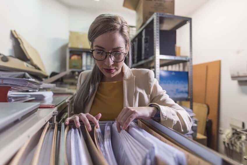 A woman wearing glasses and a beige blazer looks through files in a filing cabinet inside a storage room filled with shelves and stacks of papers.