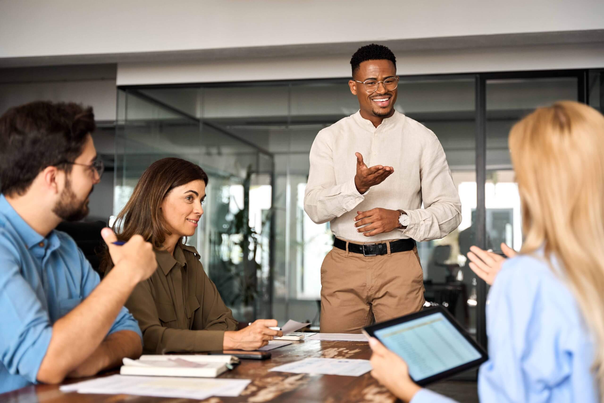 A man stands and gestures while speaking to three colleagues seated at a conference table, suggesting a team meeting or discussion in a modern office setting.