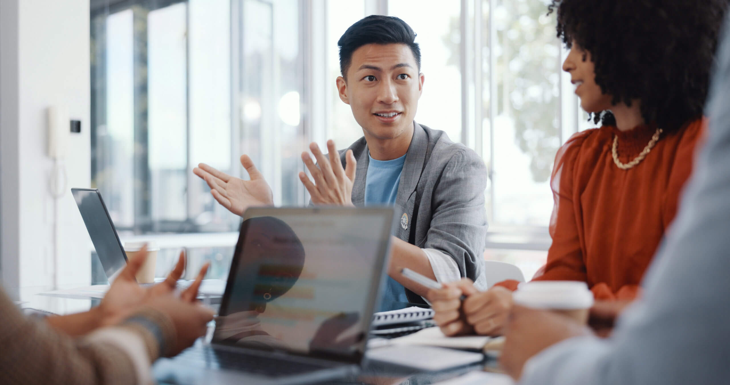 Four people sit around a table in a bright office, having a discussion. One person is speaking and gesturing with open hands, while others listen. A laptop and notebooks are on the table.