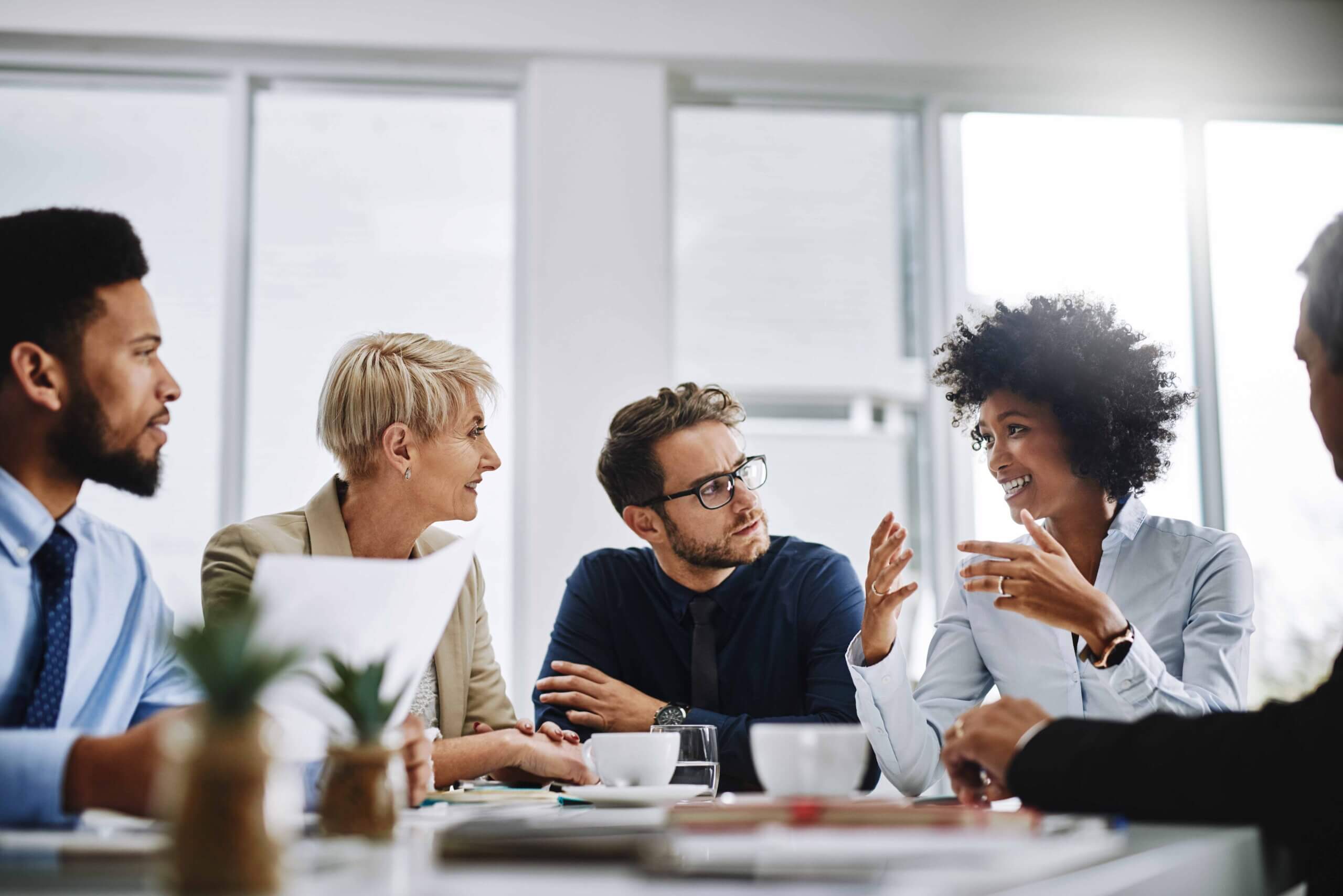 A diverse group of five professionals, including a registered agent, sits around a table in a bright office, engaged in discussion. One woman gestures animatedly while others listen attentively, with coffee cups and documents on the table.