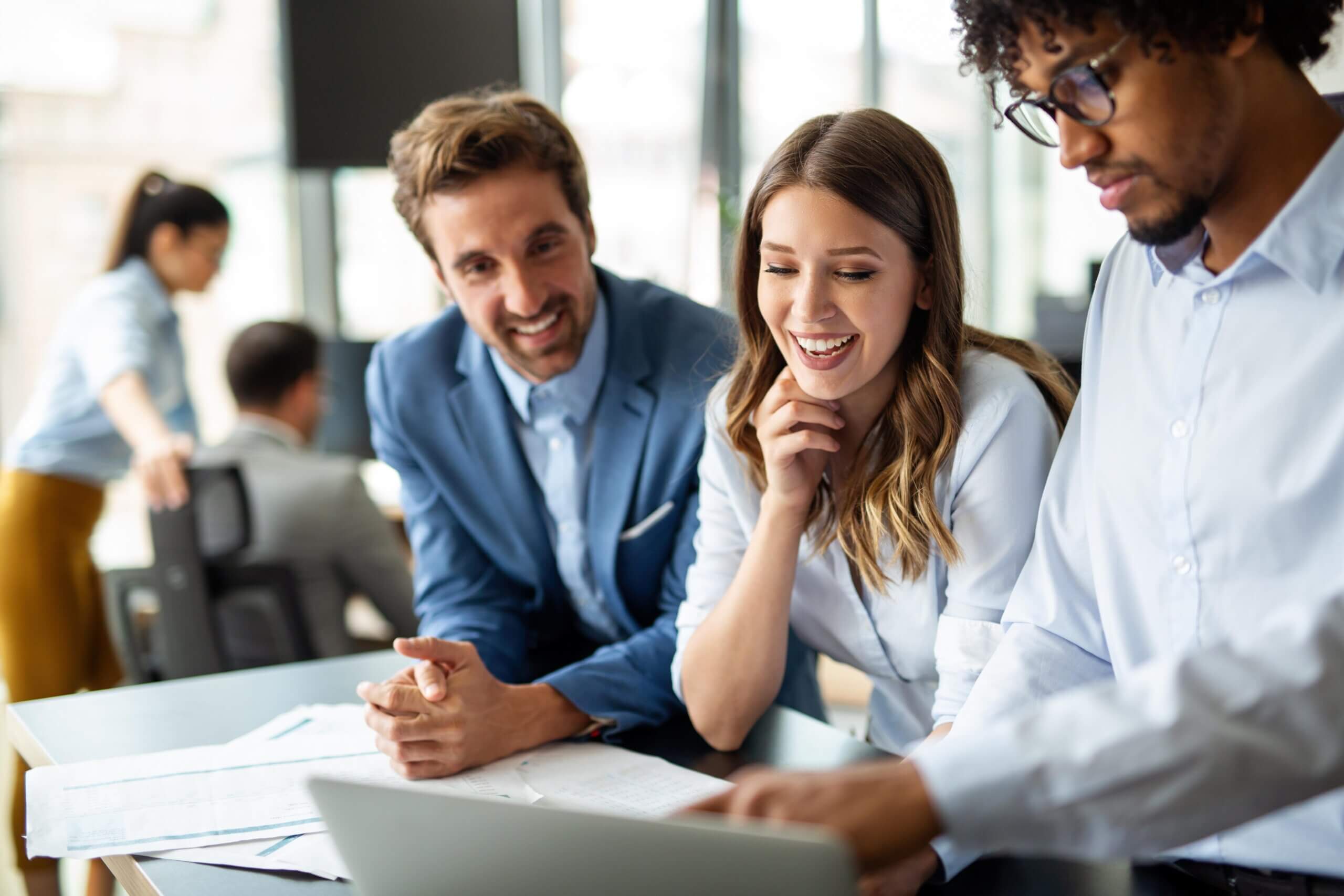 Three colleagues, two men and one woman, smile and look at a laptop screen together in a bright, modern office setting. Two other people are working in the background.
