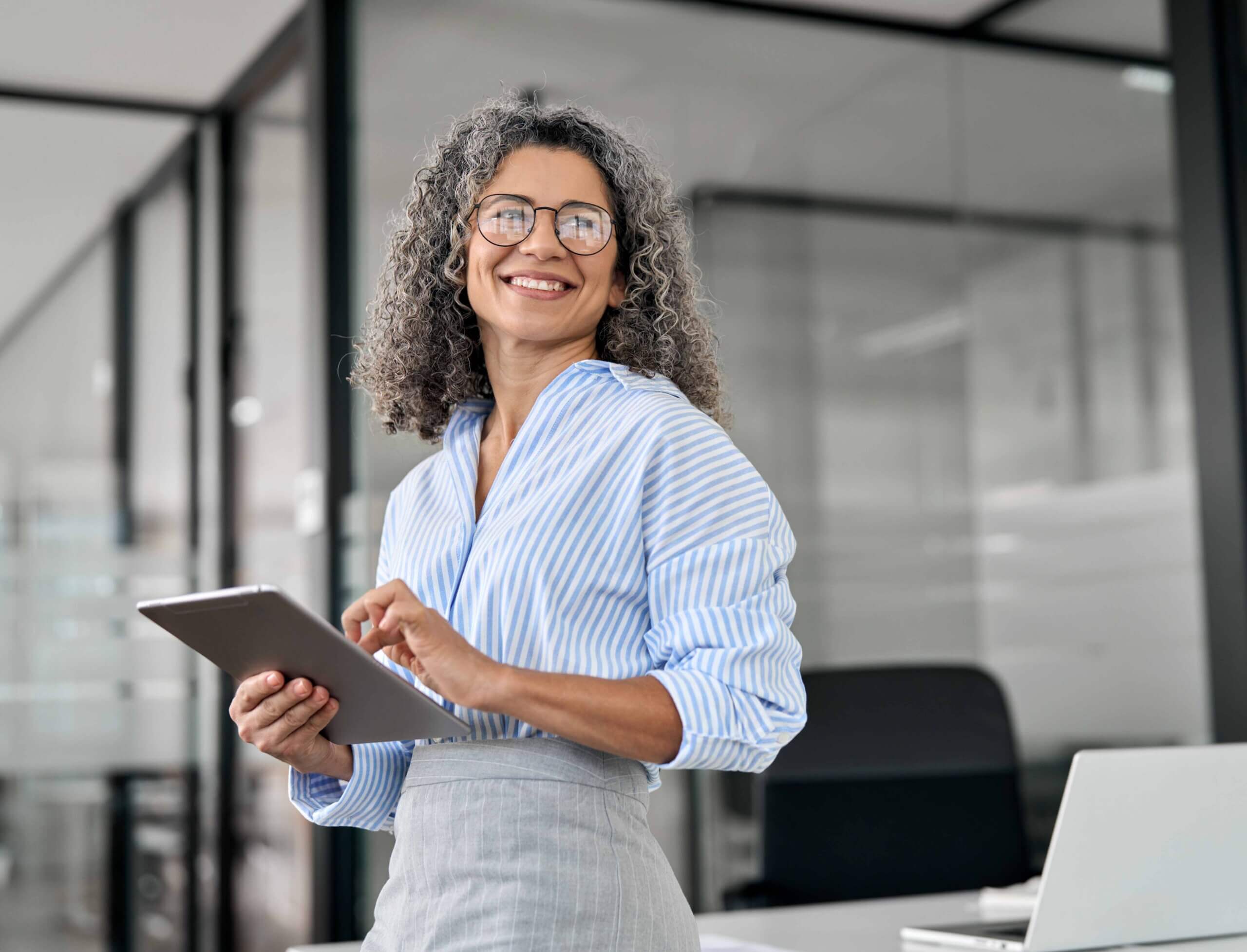 A smiling woman with curly gray hair, wearing glasses and a striped shirt, stands in a modern office holding a tablet. A laptop and office furniture are visible in the background.