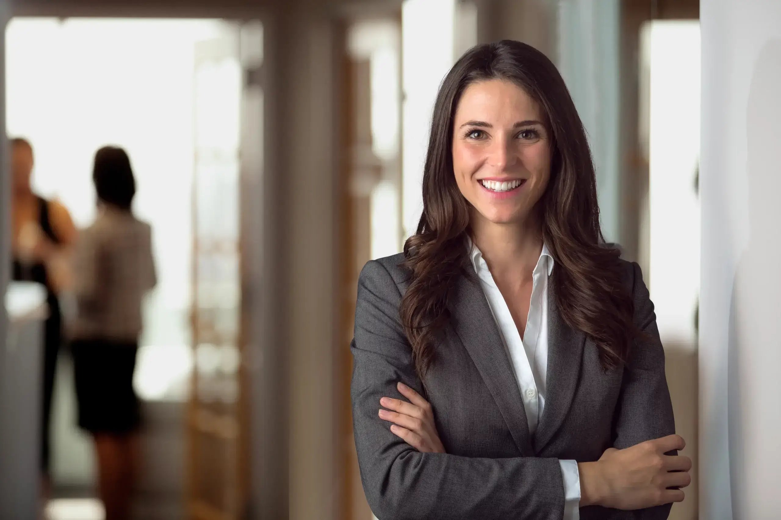 A woman in a gray blazer and white shirt stands smiling with arms crossed in a bright office hallway, while two blurred figures converse in the background.