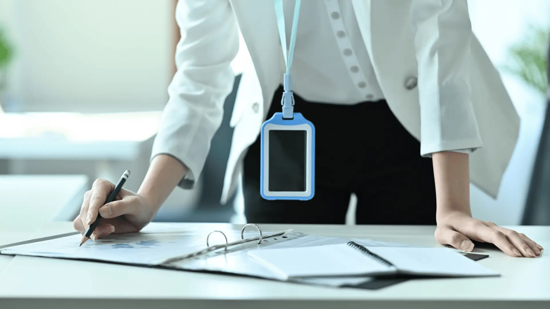 A person in a white blazer, with a blank ID badge, leans over a desk while writing on documents. Open folders and papers are spread out on the desk in a bright office setting.
