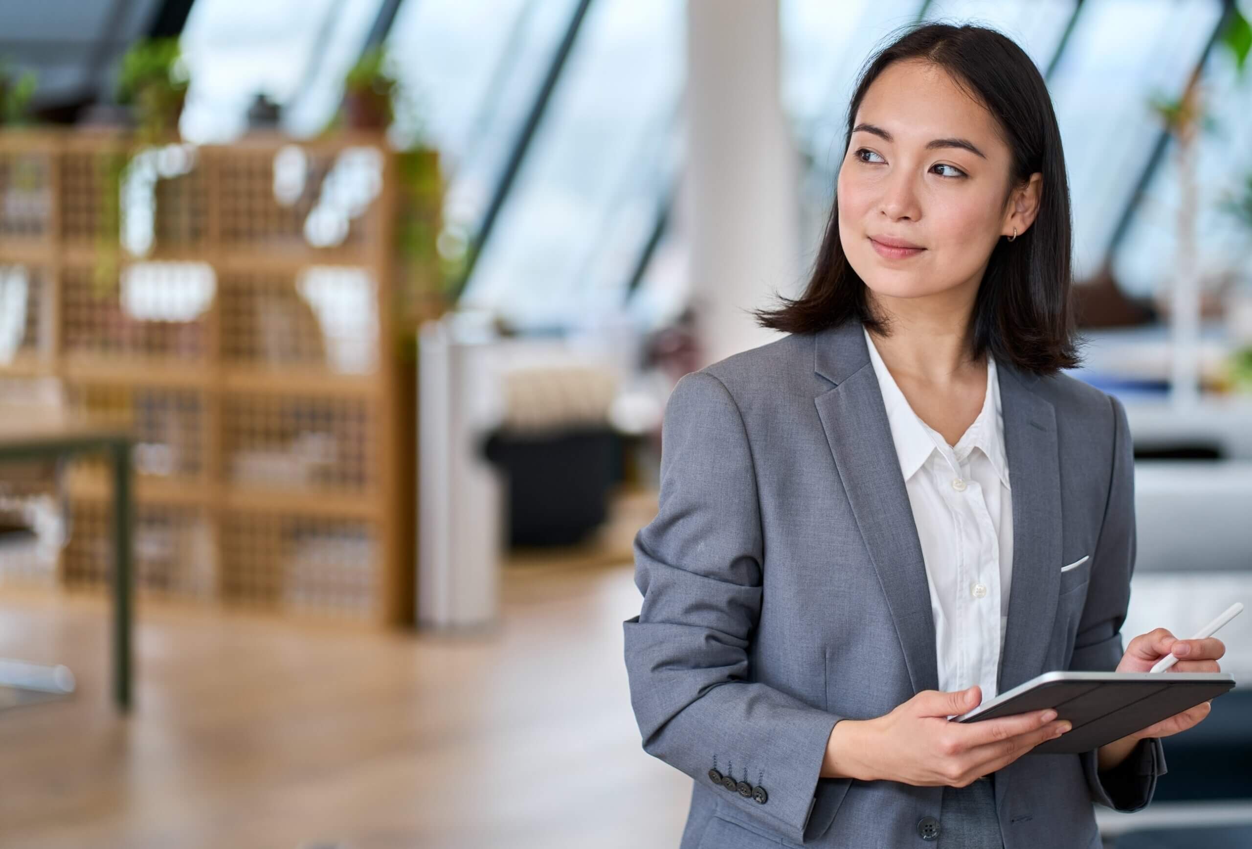 A woman in a gray suit holds a digital tablet and stylus, looking thoughtfully to the side in a modern, bright office space with large windows and shelves in the background.