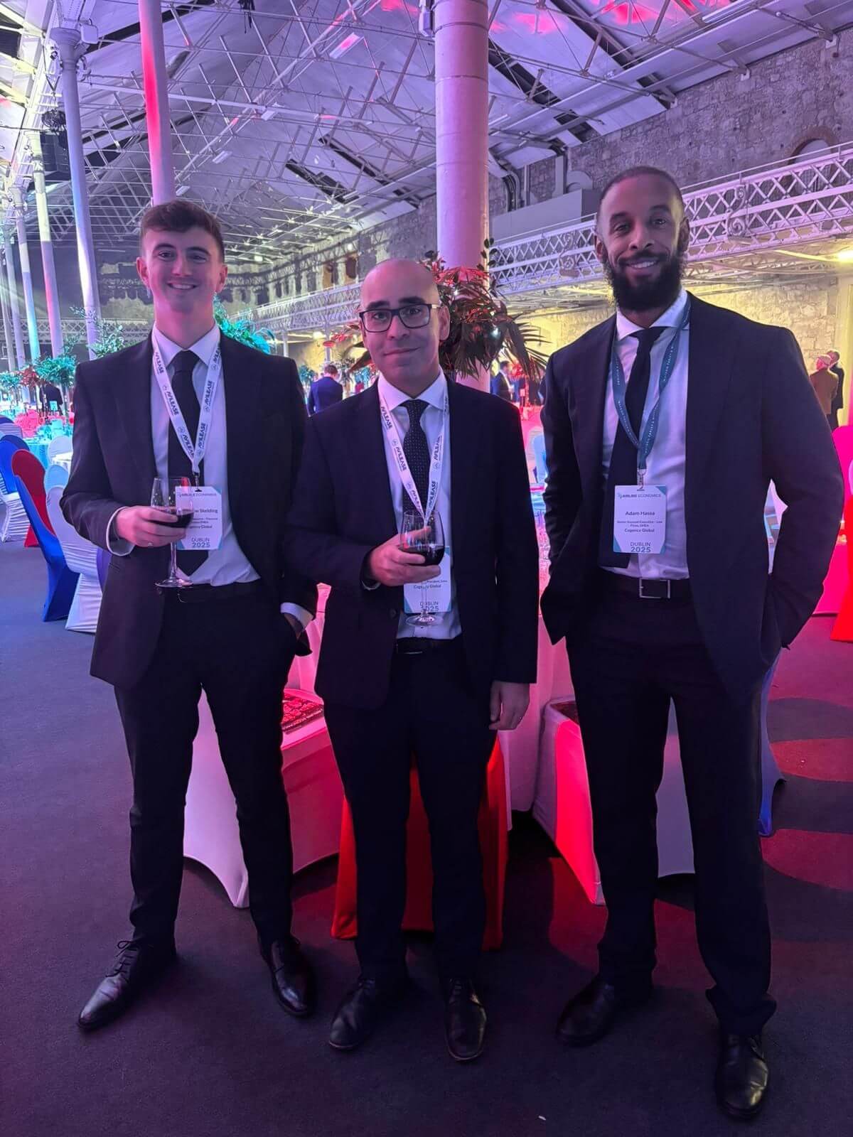 Three men in suits wearing conference badges stand indoors at an event, smiling at the camera. Two of them are holding drinks. The venue is decorated with colorful lighting and flowers.