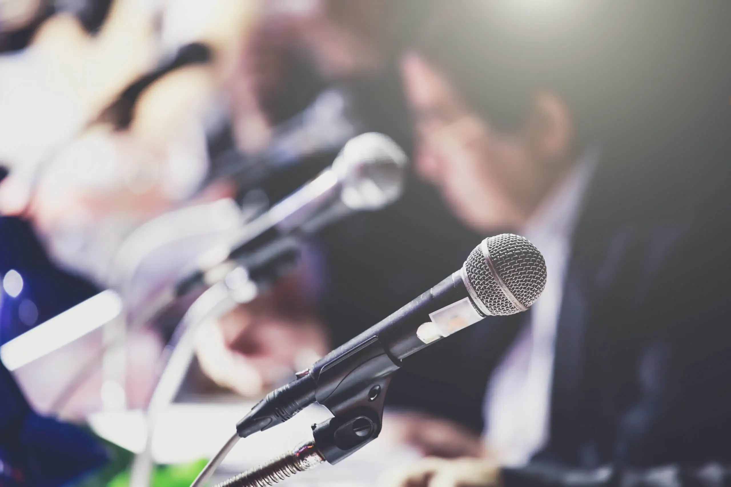 A close-up of a microphone on a stand, with several other microphones and a blurred person in the background, suggesting a press conference or public speaking event.