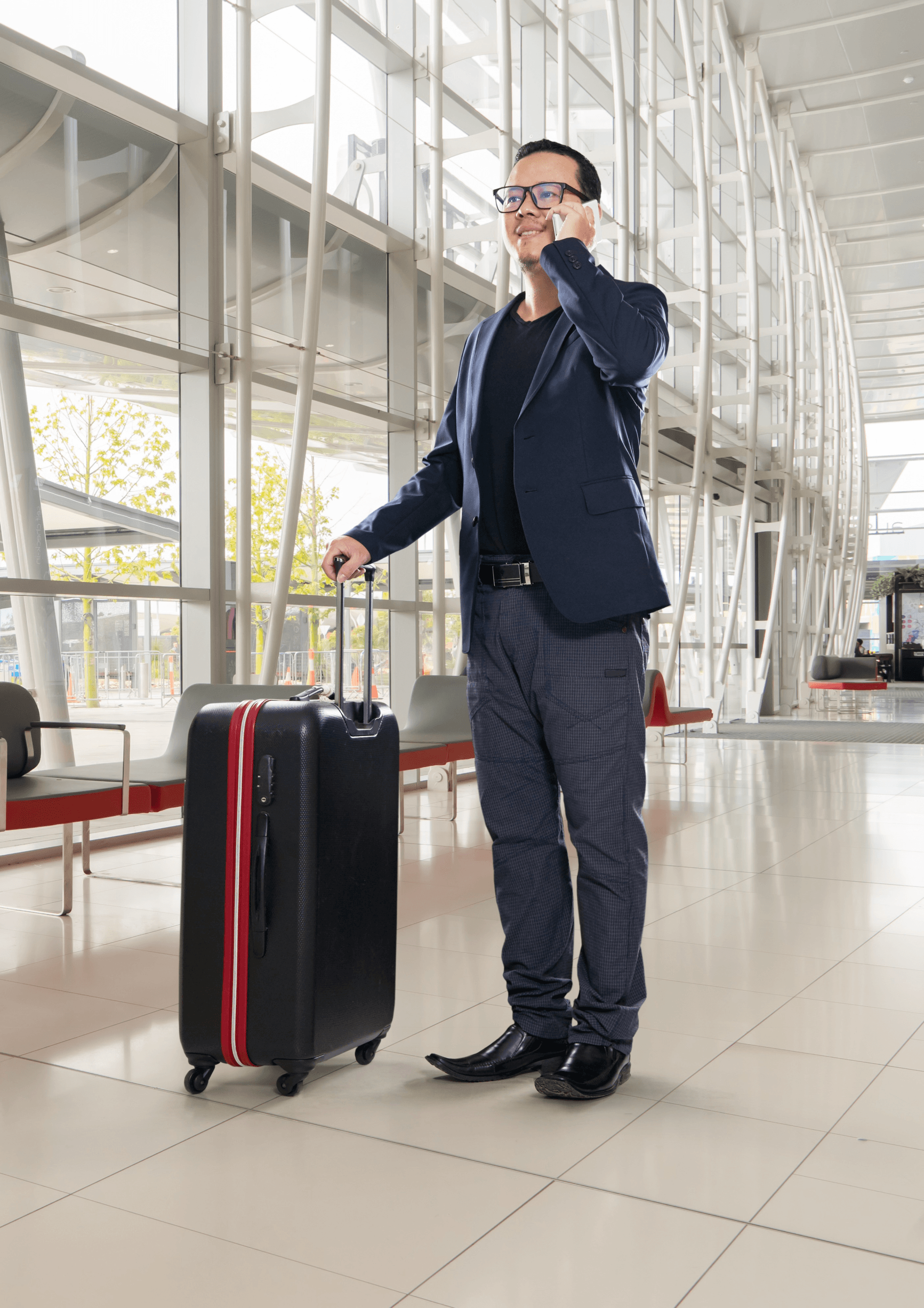 A man in a blue blazer and light sweater smiles while standing in an airport, holding a smartphone and suitcase, with a crossbody bag over his shoulder. A large blue circle is behind him.