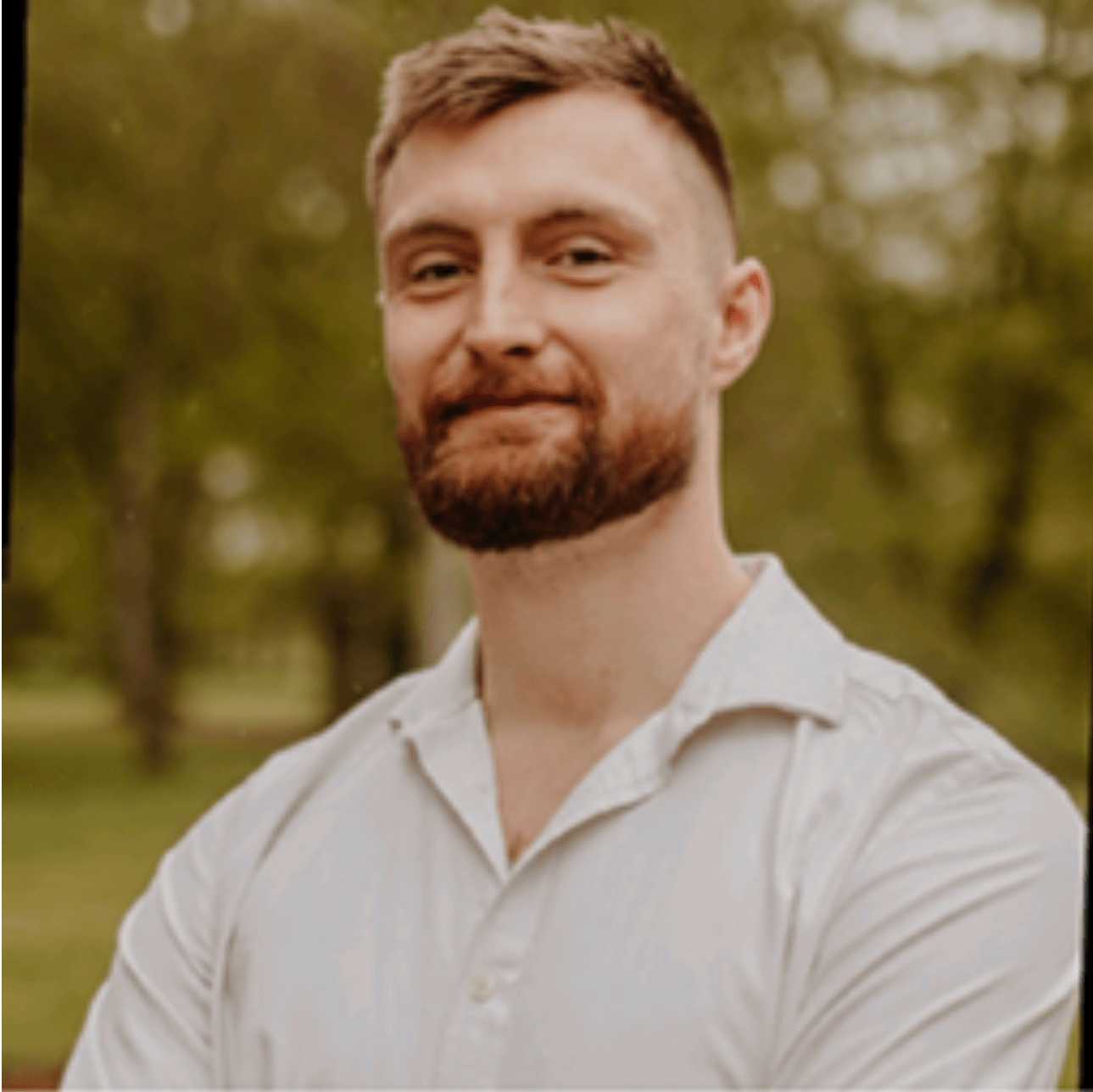 A man with short light brown hair and a beard stands outdoors, smiling slightly, wearing a white shirt. Green trees and soft natural light are in the blurred background.