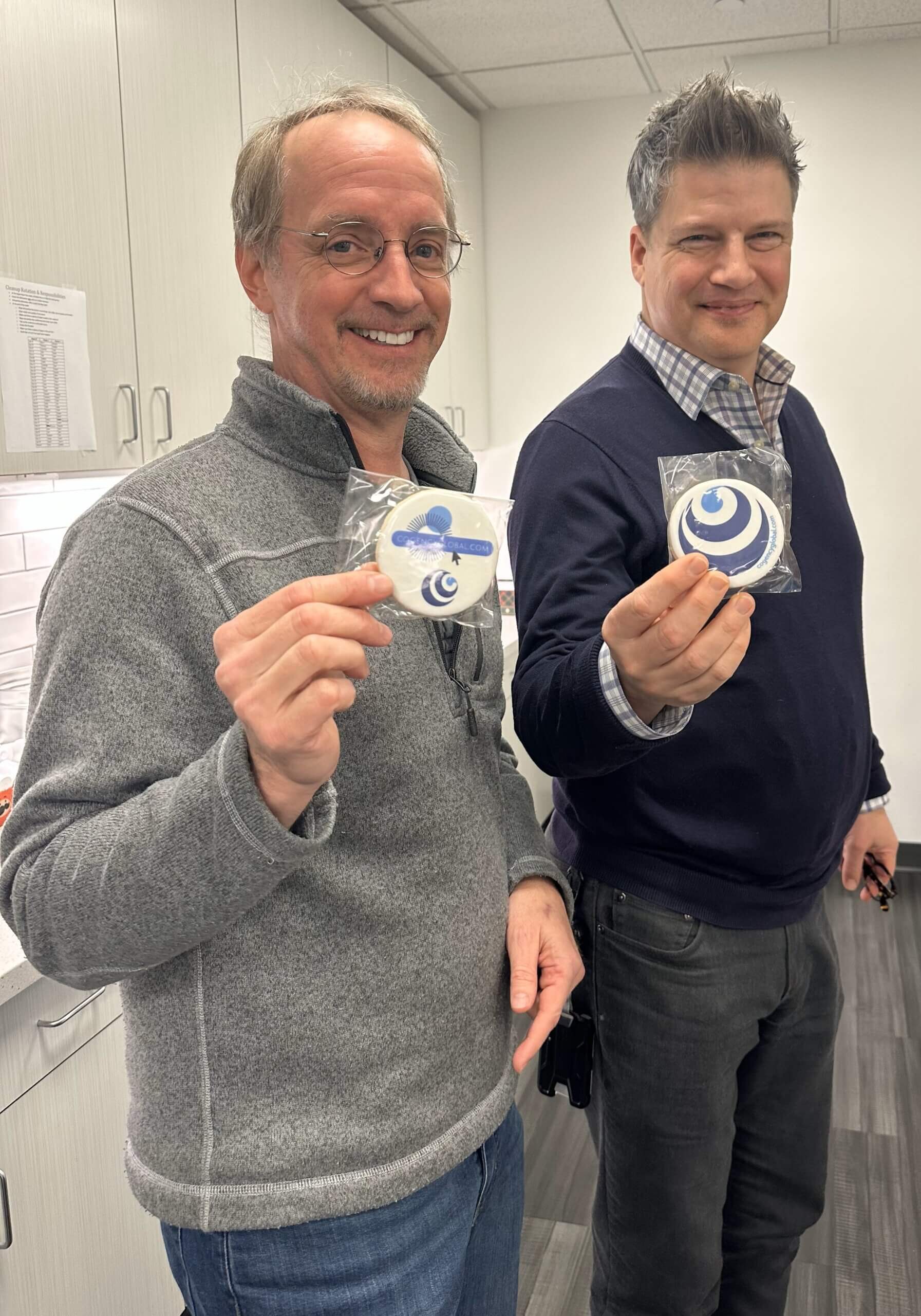 Two smiling men stand indoors, each holding a packaged cookie with a blue swirl design. They are wearing sweaters and shirts, and are in a modern office kitchen with white cabinets and gray flooring.